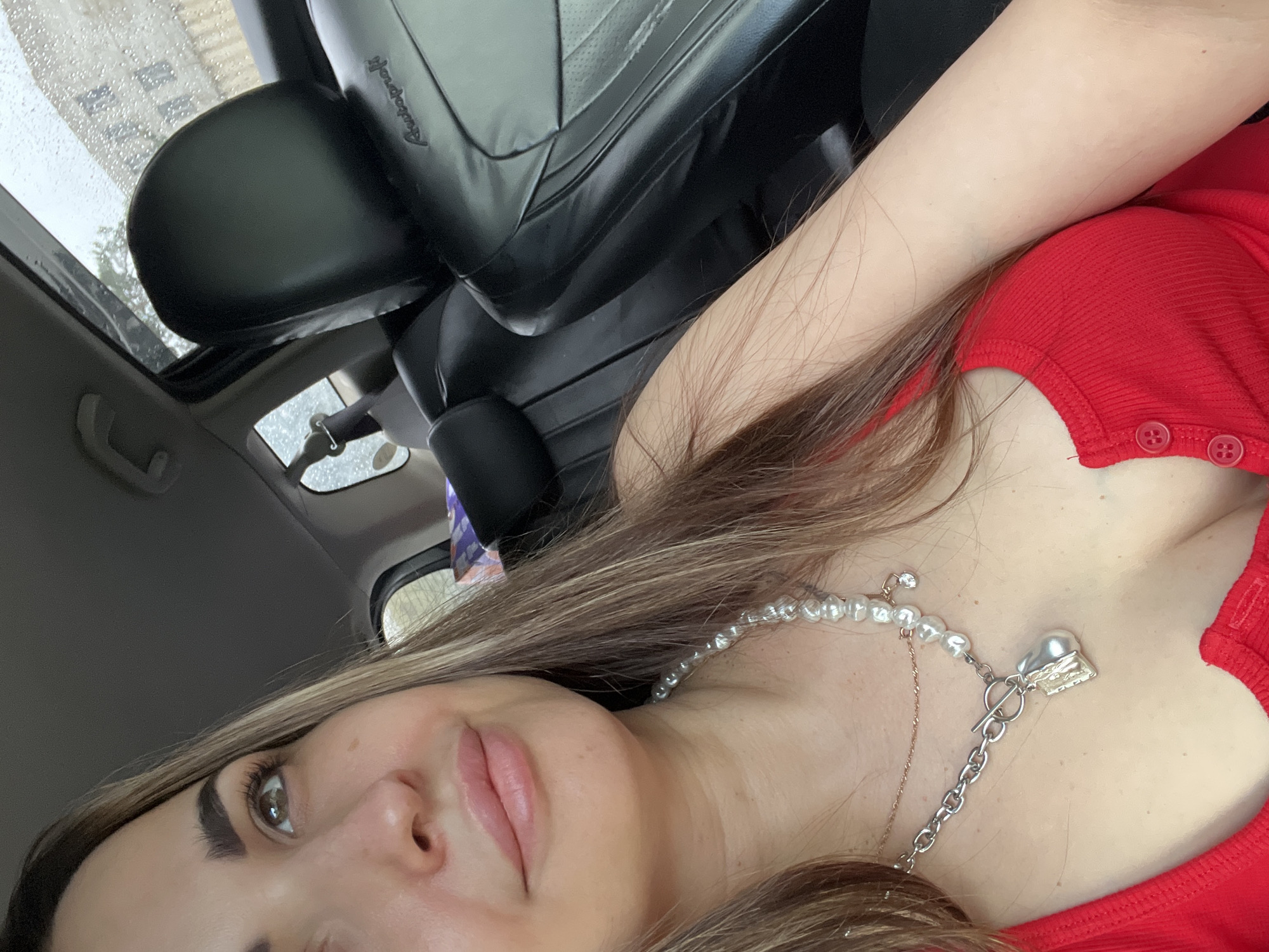 The image features a close-up, tilted view of a woman inside a car during daytime. Her face, with one eye and part of her mouth visible, has a neutral expression. She has long, straight brown hair with lighter highlights, draped over her shoulder. She is wearing a red, ribbed top with three small red buttons visible on the chest.
Around her neck, she has a layered arrangement of necklaces. The most prominent is a pearl necklace with a silver-colored toggle clasp and a large, heart-shaped pendant. A thinner, gold-colored chain is also visible, along with a small, sparkling charm attached to the pearl necklace.
The background shows the interior of a car, including a black leather seat and headrest. The word "Autoproff" is discernible on the side of the headrest. Above, a window or sunroof shows raindrops on the glass, with a blurred building in the background, indicating it is likely raining or has recently rained. The lighting suggests it is daytime. The composition suggests a selfie taken from a slightly low angle.