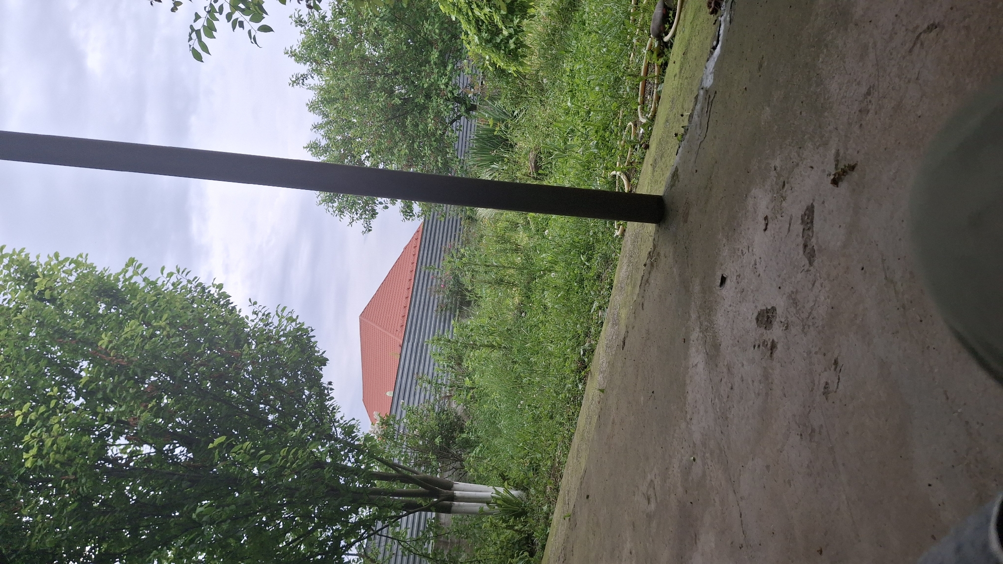 The image shows a ground-level view from what appears to be a concrete patio or sidewalk. Vegetation, including grass and trees with green foliage, is visible in the background. Part of a building with a red roof is seen through the trees. A dark, rectangular beam or post is positioned diagonally across the frame. The sky is overcast. The location appears to be outdoors, possibly a residential area. The image is rotated, but this does not prevent the recognition of key objects and details. There is no information to verify the location as Shali, Russia.