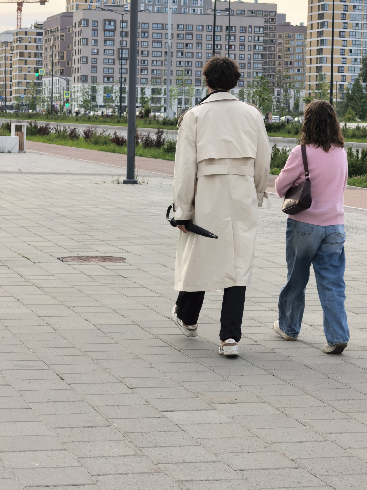 Two people, a man and a woman, are walking away from the camera on a paved sidewalk. The man is wearing a light-colored trench coat, dark pants, and white and brown sneakers. He is holding a black umbrella in his right hand. The woman is wearing a pink long-sleeved shirt, blue jeans, and white sneakers. She has a dark brown purse slung over her left shoulder.
In the background, there are several tall, modern apartment buildings with many windows. Some of the buildings are light yellow and cream, while others are gray. A street with trees and green spaces runs between the buildings and the sidewalk. Traffic lights are visible, with one showing green. A construction crane is visible on the far left side of the image, suggesting ongoing development in the area. The pavement is made of light gray rectangular tiles. The lighting suggests it is daytime, possibly late afternoon or early evening. The sky is overcast. The overall mood appears to be casual and everyday. There is no visible text in the image.
