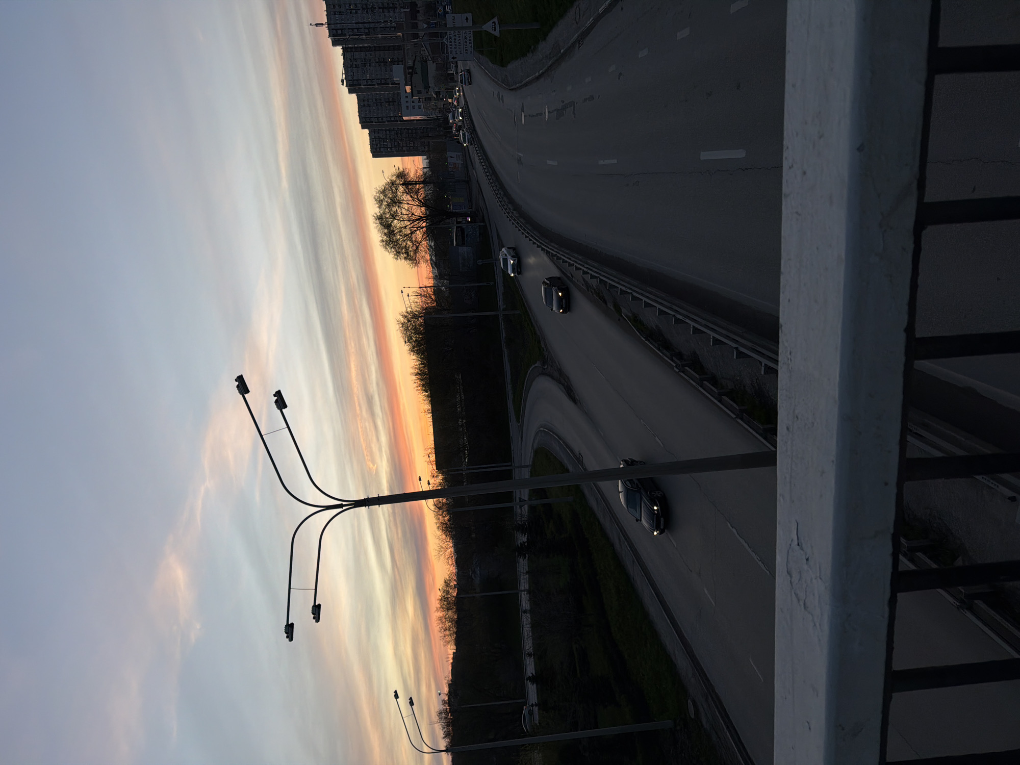 An elevated view of an urban scene at dusk, likely in Rostov-on-Don, Russia, features a multi-lane highway curving through the frame. Several cars are actively driving on the roadway. A line of tall buildings is present in the background. A cluster of trees sits alongside the road. A large, modern street lamp is in the foreground to the left. The sky exhibits a gradient of colors, transitioning from light blue to orange. A railing of a bridge is visible on the right side of the frame, suggesting the photo was taken from an elevated pedestrian crossing.