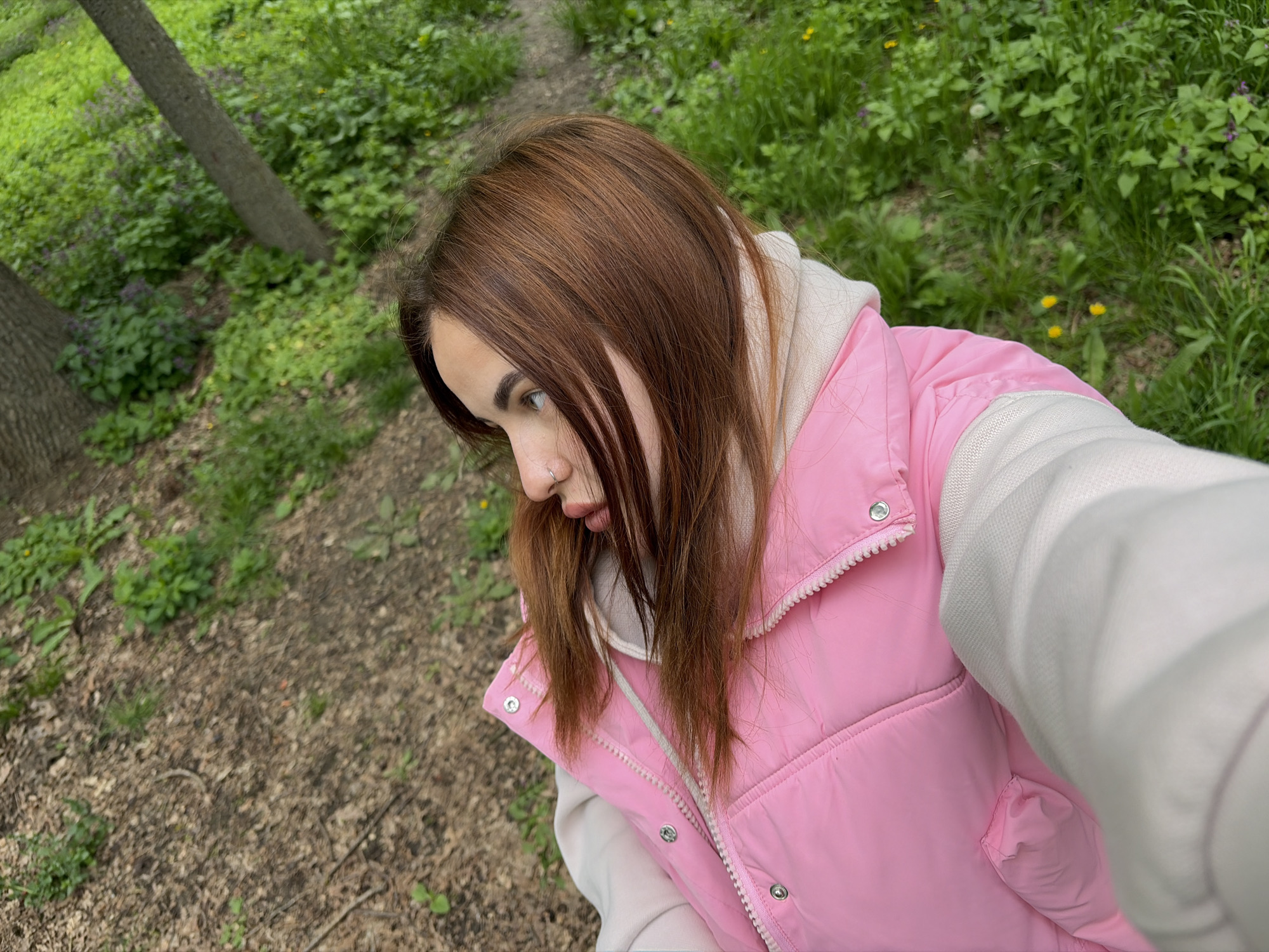 A young woman stands on a path in a wooded area. The path is covered in brown leaves and some sparse green vegetation. The woods are dense with green vegetation. The woman is wearing a pink puffer vest over a white hoodie. Her brown hair hangs loosely around her face. She is looking to her left. The location is Krasnodar, Russia.