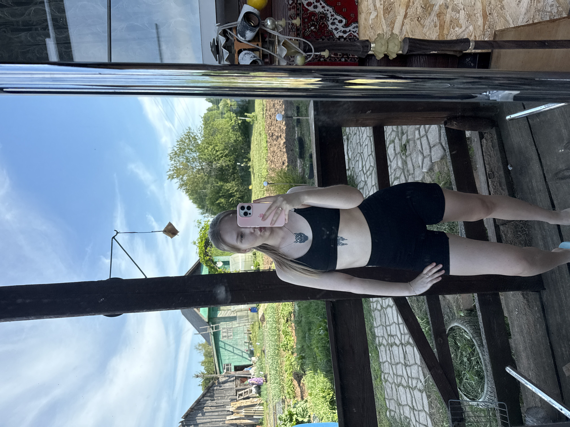 A person is taking a selfie in a mirror, in Unknown, Russia. The person is wearing a black sports bra and black shorts. They are holding a pink phone in their right hand. The mirror is reflecting a rural scene with a house, garden, and trees. The mirror is set up on a wooden structure that appears to be part of a veranda or gazebo. The ground below is paved with stone. Various objects are visible on a shelf above the mirror, including bottles, a rug, and a decorative wooden item.