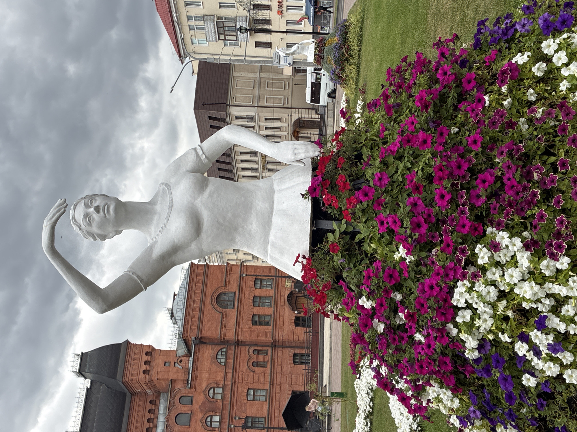A white statue of a female dancer, adorned with a skirt of colorful petunias and red flowers, stands in the foreground. Behind the statue, a red brick building with a dark roof and arched windows is visible, to the left of it, a lighter colored building with multiple floors and a red roof can be seen. Further back, a white van is parked in front of a beige building. The sky above is overcast with dark clouds. This is located in Mogilev, Belarus, specifically in the central square.