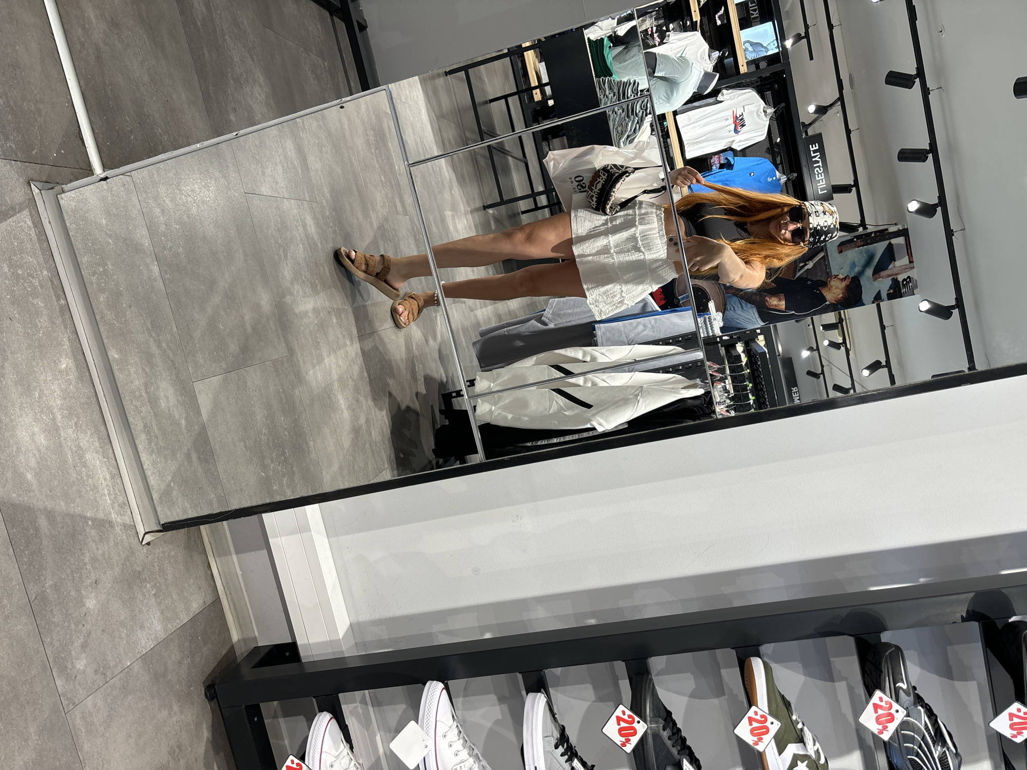 The image shows a person standing in front of a large mirror in what appears to be a retail store. The person is reflected in the mirror, wearing a white skirt, a dark top, sunglasses, and a patterned headscarf. They are also carrying a small, patterned handbag. In the background, clothing racks with various garments, including t-shirts with brand logos like "NIKE," are visible. The floor is tiled with large gray tiles.
The reflection in the mirror also shows shelves filled with shoes, some with "20%" price tags visible. The lighting in the store appears to be artificial, with spotlights directed at the displays. The overall scene suggests a shopping environment, possibly a clothing or shoe store. There is no indication of the time of day or weather outside. The location context provided is Budva, Montenegro. The person's posture and the way they are holding their hair suggest they are posing for a photo or checking their appearance.
