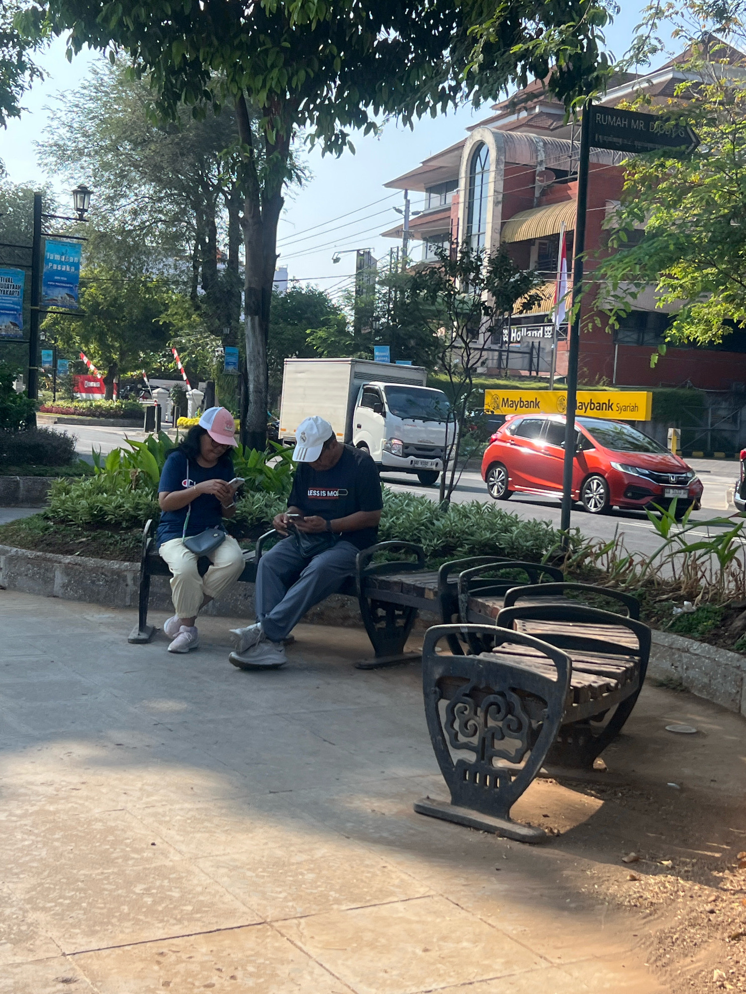 This is a horizontal, eye-level, medium shot of a street scene in the city of Yogyakarta, Indonesia, on a sunny day. In the foreground, two people are sitting on a dark metal park bench. The person on the left, wearing a navy blue shirt, cream-colored pants, a pink and white baseball cap, and pink sneakers, is looking down at a smartphone in their hands. A dark gray purse is hanging from their left shoulder. The person on the right, wearing a dark t-shirt with white lettering, dark gray pants, and a white baseball cap, is also looking down at a smartphone. Both individuals appear to be focused on their phones and are not interacting with each other.
Behind the bench, there is a decorative planter box filled with green foliage. To the right of this planter, there are two more park benches, also made of dark metal with wooden slats, positioned facing away from the viewer.
In the middle ground, a road is visible with a red Honda Jazz parked in the right lane. The car is a vibrant red color and is reflecting sunlight. Across the road, a white truck is visible. A yellow banner with black lettering that reads "Maybank Syariah" is prominently displayed. Behind the banner, a multi-story building with a reddish-brown facade and a distinctive arched window is visible. A signpost with black lettering on a white background reads "RUMAH MR. DIDI 6". Several trees with green leaves and shadows are scattered throughout the scene.
In the background, a clear blue sky is visible. Further in the distance, there are more buildings and trees, suggesting a busy urban environment. The overall impression is that of a public space with people casually using their phones while sitting on a bench, with cars and buildings forming the backdrop.