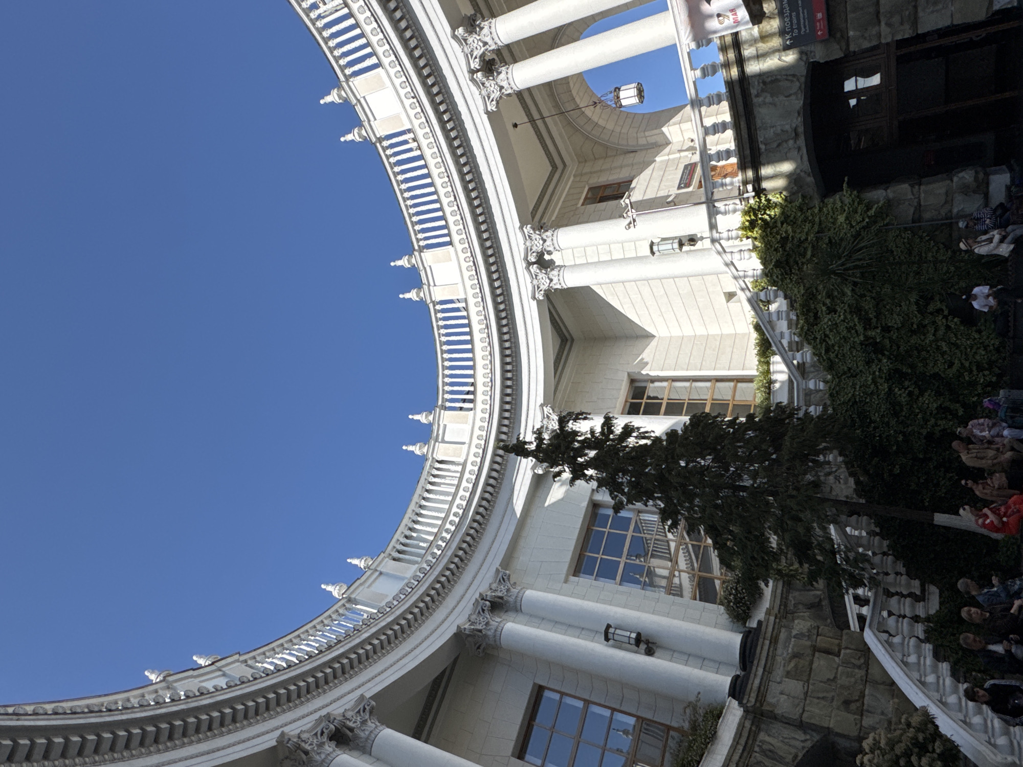 The image shows the architectural details of the Sochi railway station in Sochi, Russia. The shot is taken from a low angle, looking up at the station's circular, colonnaded facade against a clear blue sky. The building's white columns and ornate details are prominent. A tree and some landscaping elements are visible in the foreground, partially obscuring the base of the building. There are people situated near the entrance.