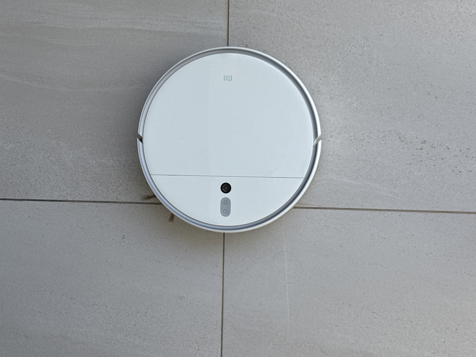 An overhead shot shows a white, circular robot vacuum cleaner positioned on a light gray tiled floor. The vacuum has a metallic silver trim around its outer edge. On the top surface of the vacuum, there are two circular buttons, one with a power icon, and a black circular sensor. The Xiaomi logo, an “MI” in a square, is visible in the upper right quadrant of the vacuum's surface. The floor is composed of large, rectangular tiles with visible grout lines. No people are present in the scene. The image does not provide any information to determine its location within Paphos Municipality, Cyprus.