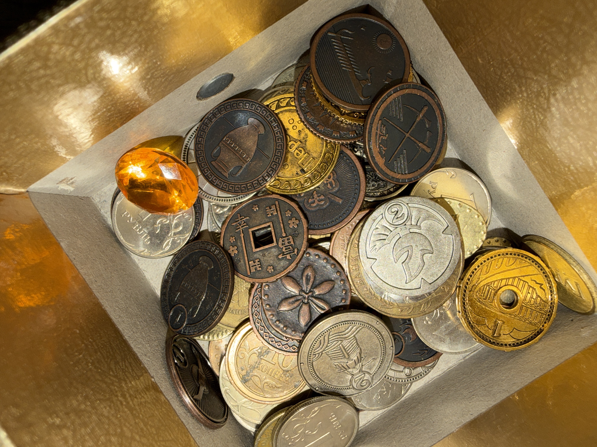 The image shows a variety of coins and a faceted amber gemstone inside a container with a golden interior. The coins are of different sizes, colors (silver, gold, bronze), and designs. Many appear to be commemorative or collectible coins, featuring images of buildings, symbols, and historical figures. Some coins bear inscriptions in different languages. The amber gemstone is a yellowish-orange color and is located near the edge of the container, resting on top of several silver coins. The container itself appears to be made of cardboard or a similar material, with a beige or light gray interior lining. The overall lighting is bright and highlights the metallic surfaces of the coins and the gemstone. There is no indication of the location being Votkinsk, Russia.