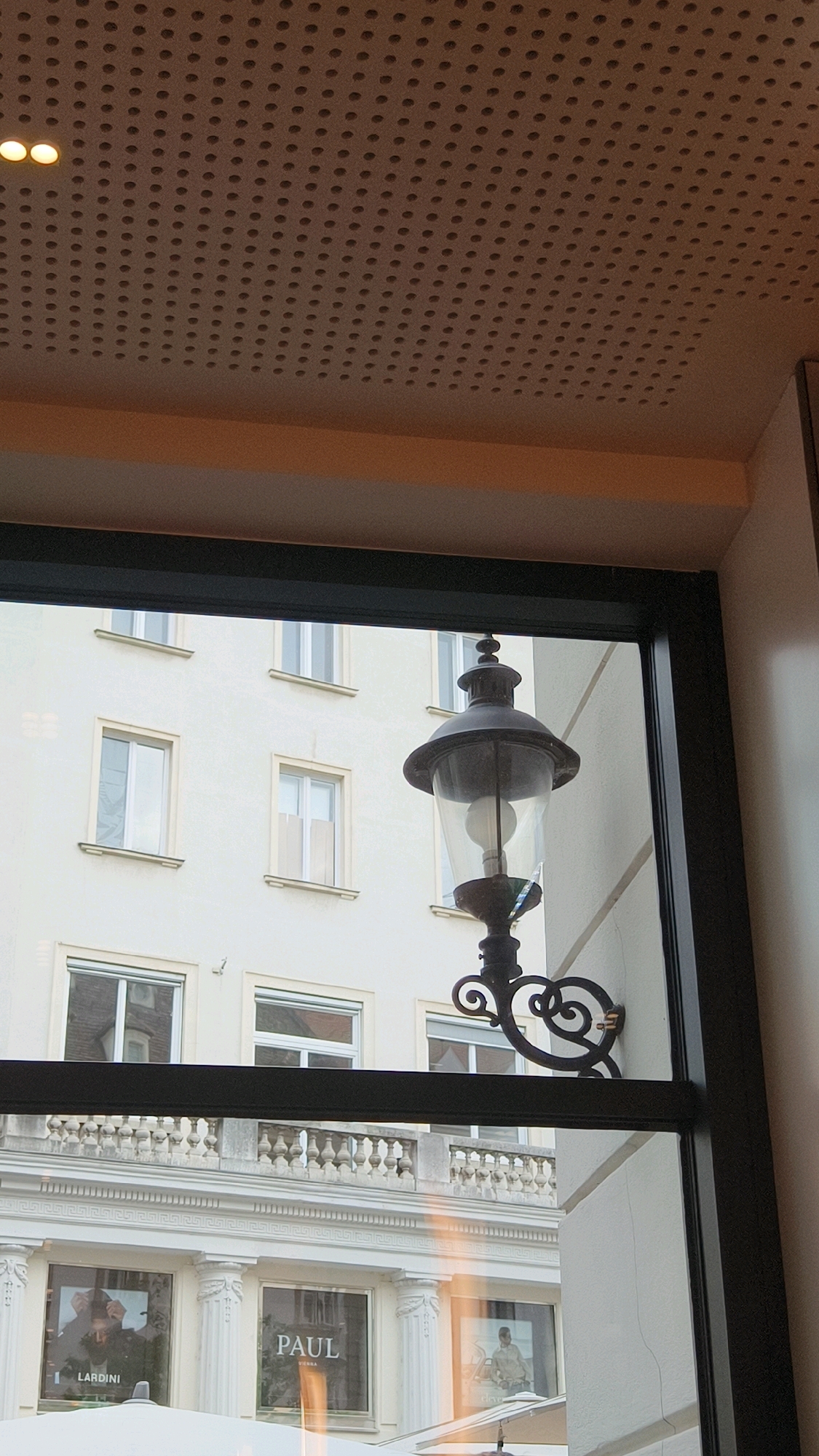 The image shows a view through a window of a building facade in Vienna, Austria, with a black framed window opening into the scene. In the foreground, a black metal lamppost is mounted on the wall to the right, with an ornate scroll design supporting its base. The lamppost itself has a dark, metallic finish and features a glass lantern with a visible light bulb inside, and a decorative finial at the top.
Beyond the lamppost, a building with a light-colored facade is visible, featuring multiple windows. The windows are rectangular with white frames and some have subtle shading or coverings inside. There is a decorative balustrade running along a level of the building above street level, which is supported by columns. Below this balustrade, the facade has architectural details including pilasters and a Greek key frieze.
At the street level, several large display windows are visible, showcasing advertisements. One window prominently displays the text "PAUL VIENNA", suggesting a store or establishment named Paul in Vienna. Another window shows an advertisement for "LARDINI". The scene appears to be an outdoor environment, likely a street in a city, given the building architecture and storefronts. The lighting suggests it could be daytime, possibly overcast or in the late afternoon, with some warm light reflecting on the facade from the right side of the image. The ceiling above the window frame shows a perforated surface with two illuminated lights, indicating an indoor space from which the photograph was taken.