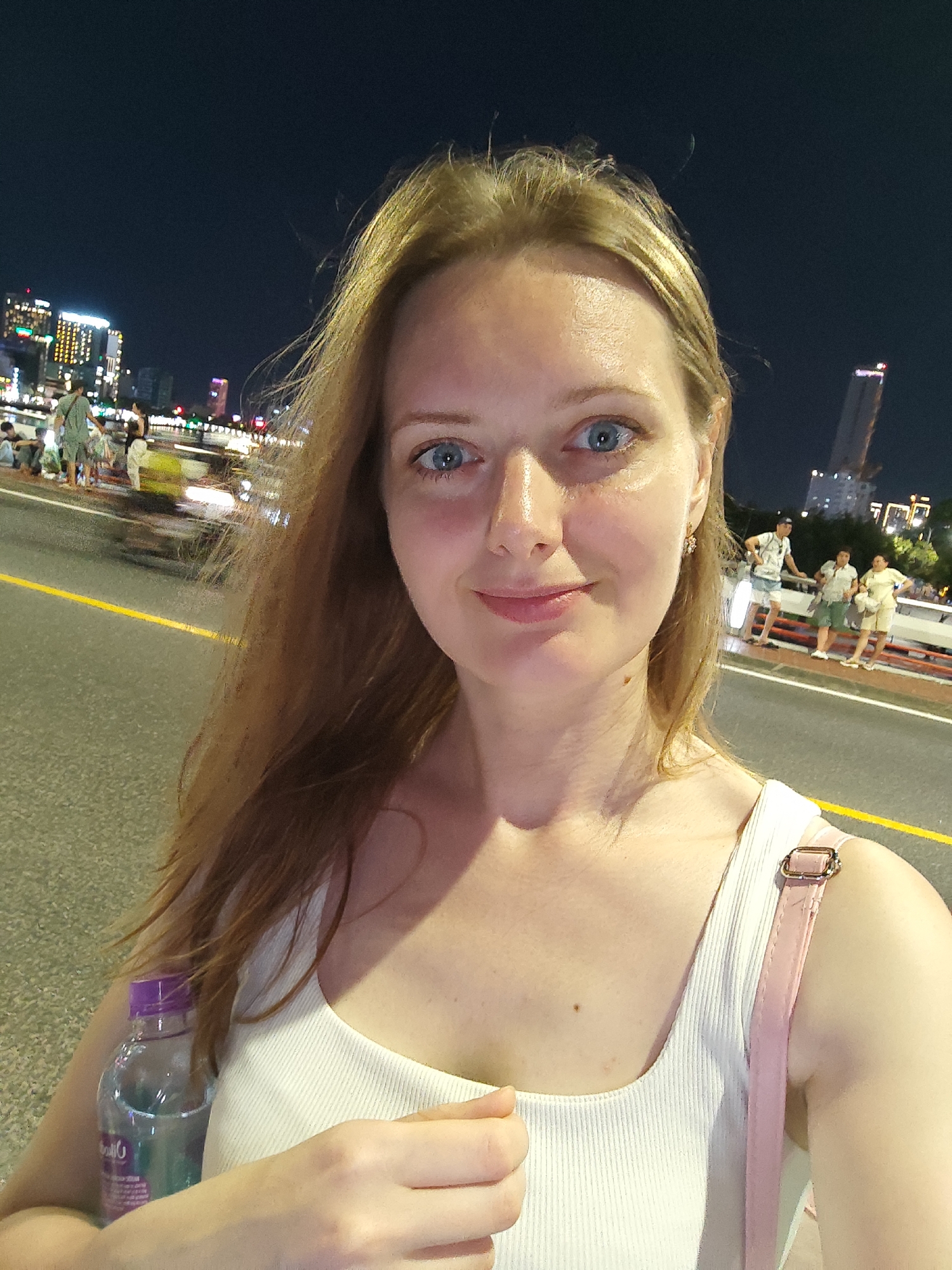 A woman with light blonde hair and blue eyes is taking a selfie on a bridge at night. She is wearing a white tank top with a light pink strap. She is holding a water bottle in her left hand. In the background, there are people walking on the bridge and buildings in the city of Đà Nẵng, Vietnam. A yellow line is painted along the side of the bridge.