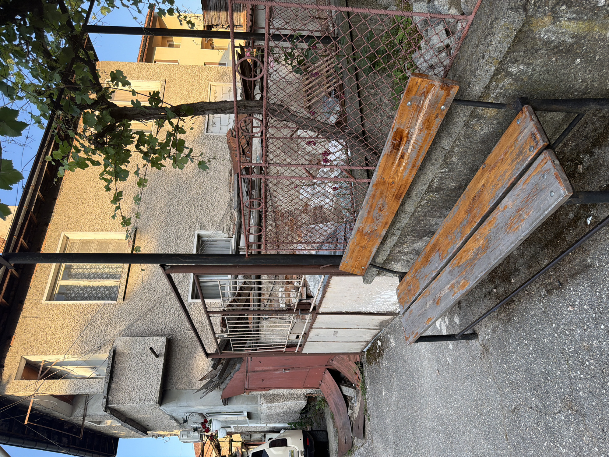 The image depicts a weathered wooden bench situated on a concrete ledge in front of a chain-link fence. Behind the fence, a building with textured walls and an overgrown vine-covered structure is visible. To the left of the building, a red metal door and debris are present, alongside a parked white vehicle. The scene appears to be an outdoor area within a residential or urban setting, possibly in Pazardzhik, Bulgaria, given the architectural style and the presence of common urban elements. The lighting suggests it is either early morning or late afternoon, with sunlight casting warm tones on the building.