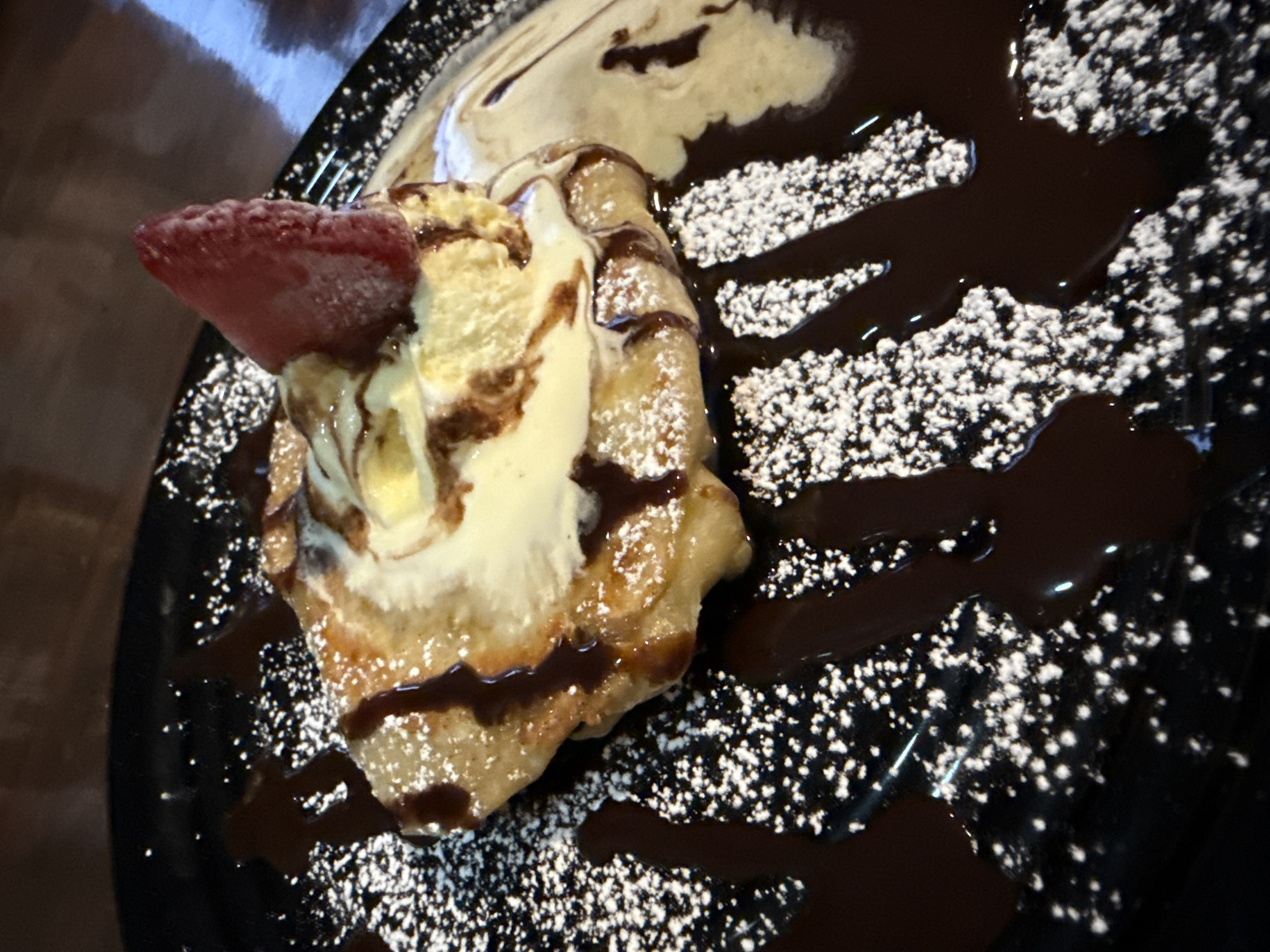 A close-up shot depicts a dessert served on a black plate. The dessert comprises a pastry topped with a scoop of ice cream, drizzled with chocolate sauce, and garnished with a sliced strawberry. The plate is decorated with powdered sugar and chocolate syrup. The setting suggests it could be a restaurant or cafe. While the image itself doesn't explicitly pinpoint the location in Puebla, Mexico, the style of dessert and plating might be characteristic of culinary offerings found in the region.