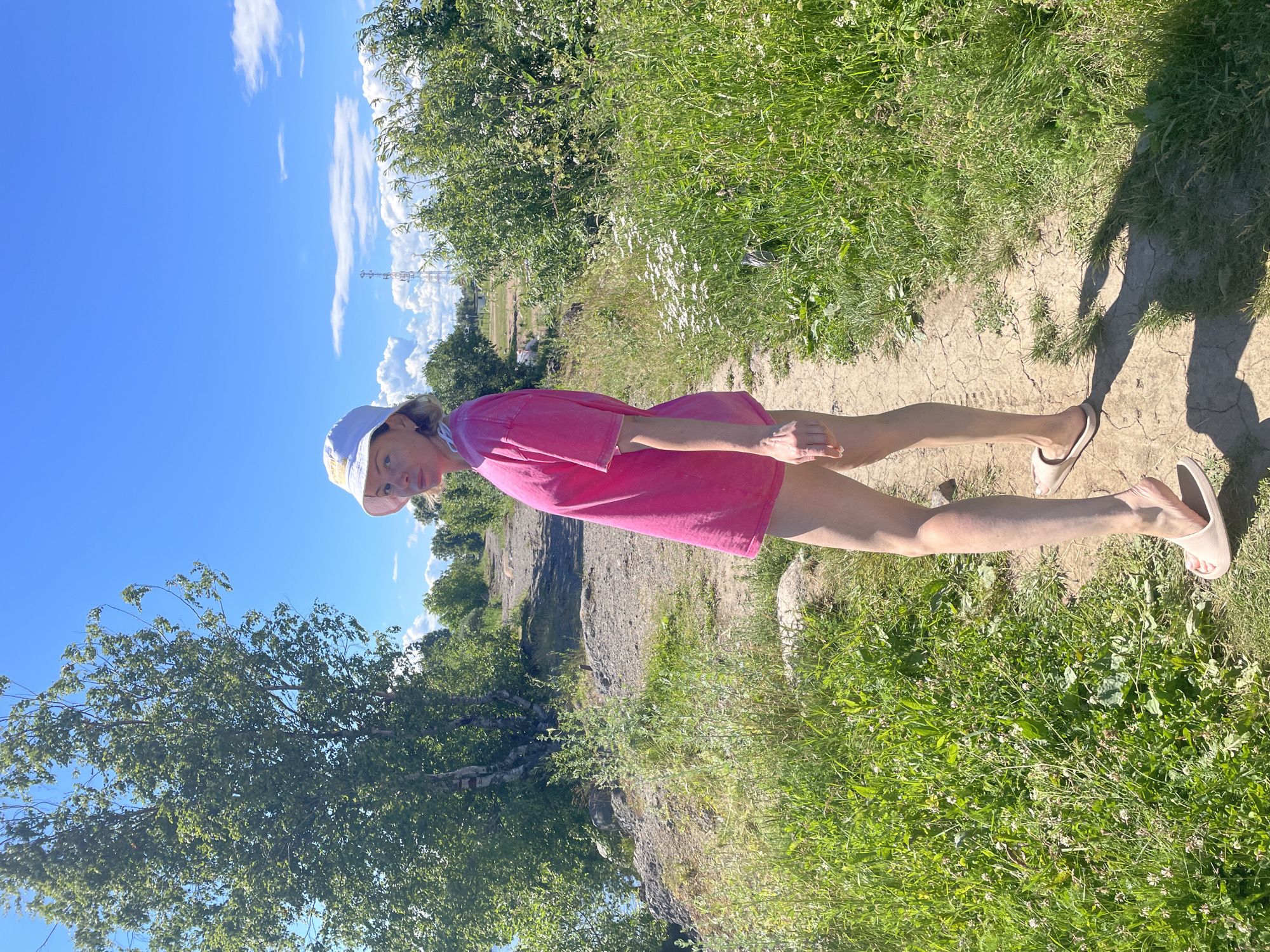 A young woman with blonde hair, wearing a pink t-shirt and a white cap, stands on a path in a grassy area with trees. She is facing towards the viewer, with her head tilted slightly. The sun is shining brightly, casting shadows on the ground, suggesting it is daytime. The sky is a clear blue with a few white clouds. In the background, there are more trees and a rocky, uneven landscape. A cell tower can be seen in the distance. The woman appears to be relaxed and enjoying the outdoors. The image is taken in Belozersk, Russia.