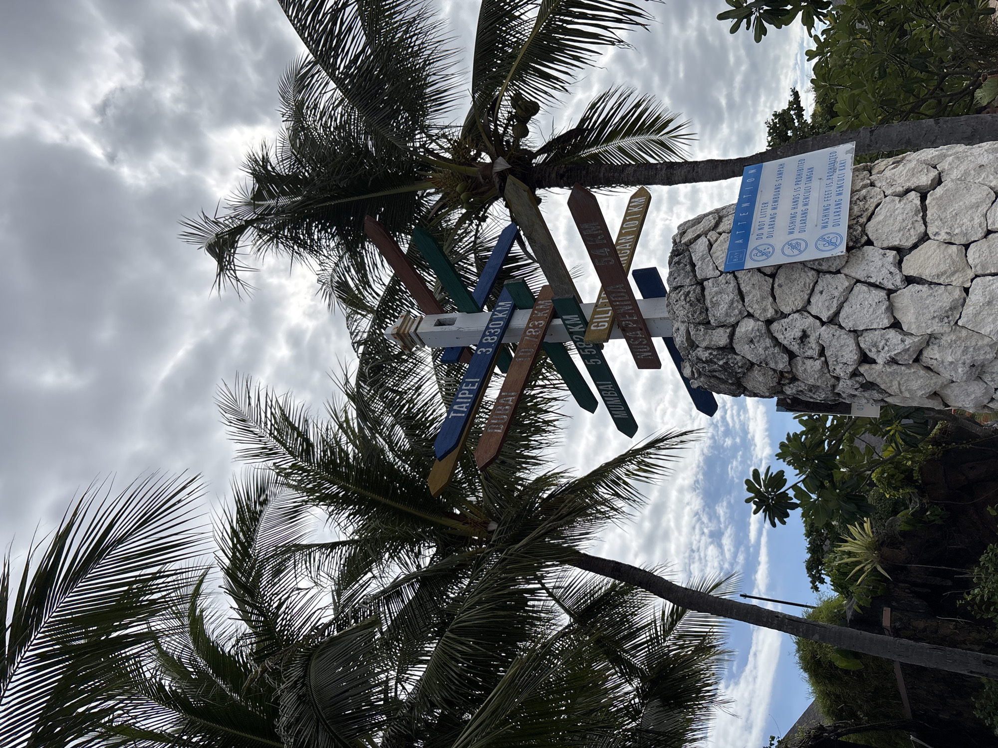 The image shows a signpost with directions and distances to various cities, including Taipei, Dubai, and Mumbai, situated in Kuta Selatan, Indonesia. The signpost is surrounded by palm trees. The signpost is made of a stone base with a blue sign indicating that littering, washing hands, and washing feet are prohibited. Several painted wooden signs are attached to the top, pointing in different directions. Some signs indicate distances to places such as Gilitrawangan, Nusa Penida, and Negara. The sky behind the trees and sign is cloudy. Vegetation and parts of buildings can be seen in the background.