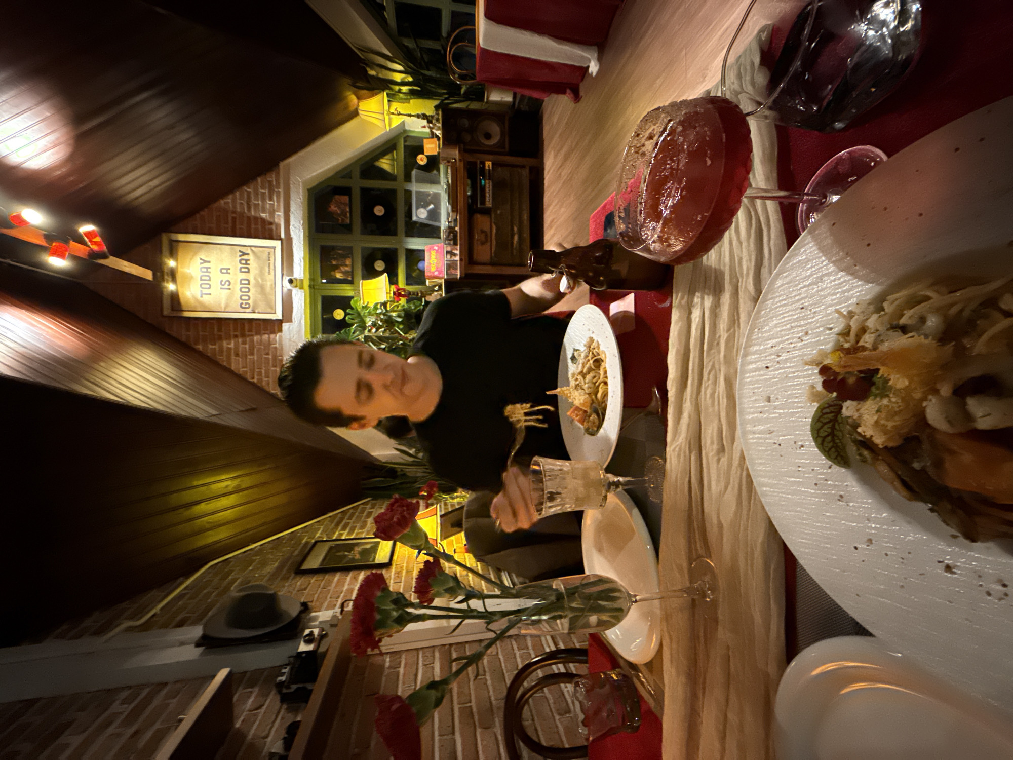 A man in a black shirt is seated at a table in a restaurant located in Lâm Viên - Đà Lạt, Vietnam. He is eating pasta with a fork. On the table is a plate with a large serving of food, a glass of red liquid, a bottle of beer, a flower vase with red carnations, and other table settings. A framed sign on the wall reads "TODAY IS A GOOD DAY." The interior is lit with warm lighting and decorated with vintage items. A hat is placed on a shelf on the brick wall.