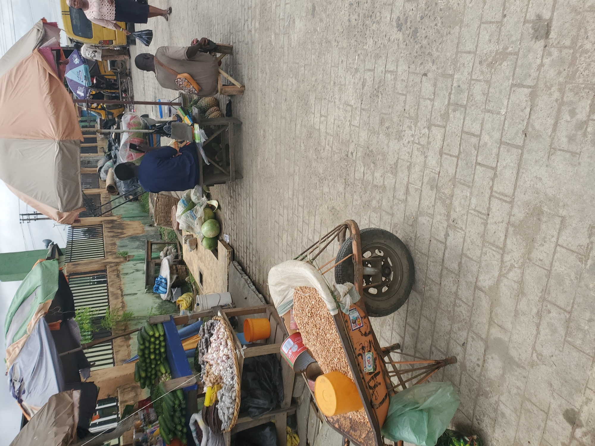 The image captures a vibrant outdoor market scene on a paved street in Lagos, Nigeria, under bright daylight. The street, made of light-colored interlocking pavers, forms the foreground and right side of the frame.
On the left side of the street, several market stalls are set up. One prominent stall is shaded by a large, light-colored umbrella. Behind this, other makeshift stalls display various goods. A person in a dark blue top is seated at one of these stalls, looking down, possibly at a phone or items on display. Further along the street, another person in a light brown shirt is seated on a low wooden stool behind a stall laden with pineapples and other produce. This person has a small patterned bag slung over their shoulder. In the background, a yellow tricycle taxi (keke Napep) is visible, with other people standing or moving around it.
Towards the far right of the scene, a woman in a light-colored blouse and dark skirt walks away from the viewer, carrying a striped bag.
Closer to the foreground, on the right side of the paved path, is a sturdy, rusty pushcart or wheelbarrow. It holds a large white sack filled with what appears to be dried beans or groundnuts, alongside orange buckets. Bunches of garlic are strung together and displayed on a wooden stand further along the left. Other produce, resembling green plantains or bananas, are also visible on various stalls. The background features weathered buildings with slatted windows and some overhead utility lines.
While much of the text is unreadable, a sticker on the pushcart displays a logo with "28" and other small, indistinct characters. No strong emotions are discernible, as people appear to be engaged in everyday market activities.