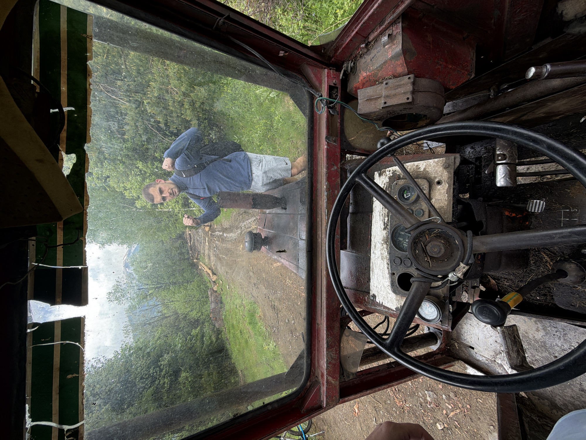 The image shows the interior of an old, red vehicle, possibly a truck or tractor, with a steering wheel and dashboard visible. The dashboard is worn and covered in dirt. Through the cracked and dirty windshield, a reflection of a man in a blue shirt and light-colored shorts is visible. The background seen through the windshield and reflected is a forest. The location is Рилски манастир, Bulgaria. The interior of the vehicle appears derelict and unused. The man is standing outside the vehicle, seemingly posing for the photo.
