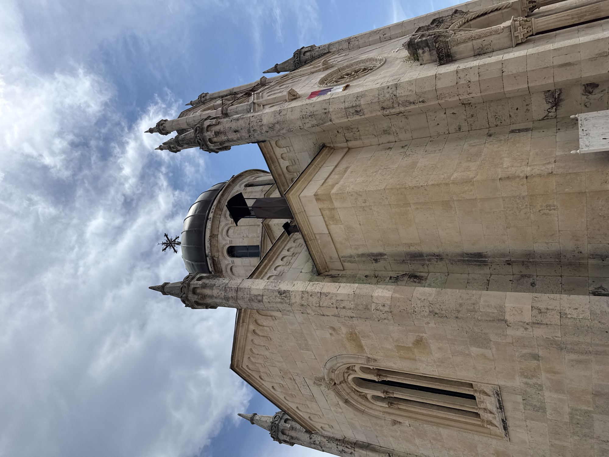 The image is a low-angle shot looking up at the facade of a stone building, likely a church, under a cloudy blue sky. The architecture appears to be classical with ornate stonework, including arched windows and decorative elements on the spires. A dark dome is visible near the top of the building, topped with a cross. A small flag, possibly of Serbia, is attached to the side of the building. The stonework is weathered, showing signs of age and exposure to the elements. The light suggests it is daytime, with diffused sunlight filtering through the clouds. The scene evokes a sense of historical grandeur and religious significance. The location is identified as Herceg Novi, Montenegro.