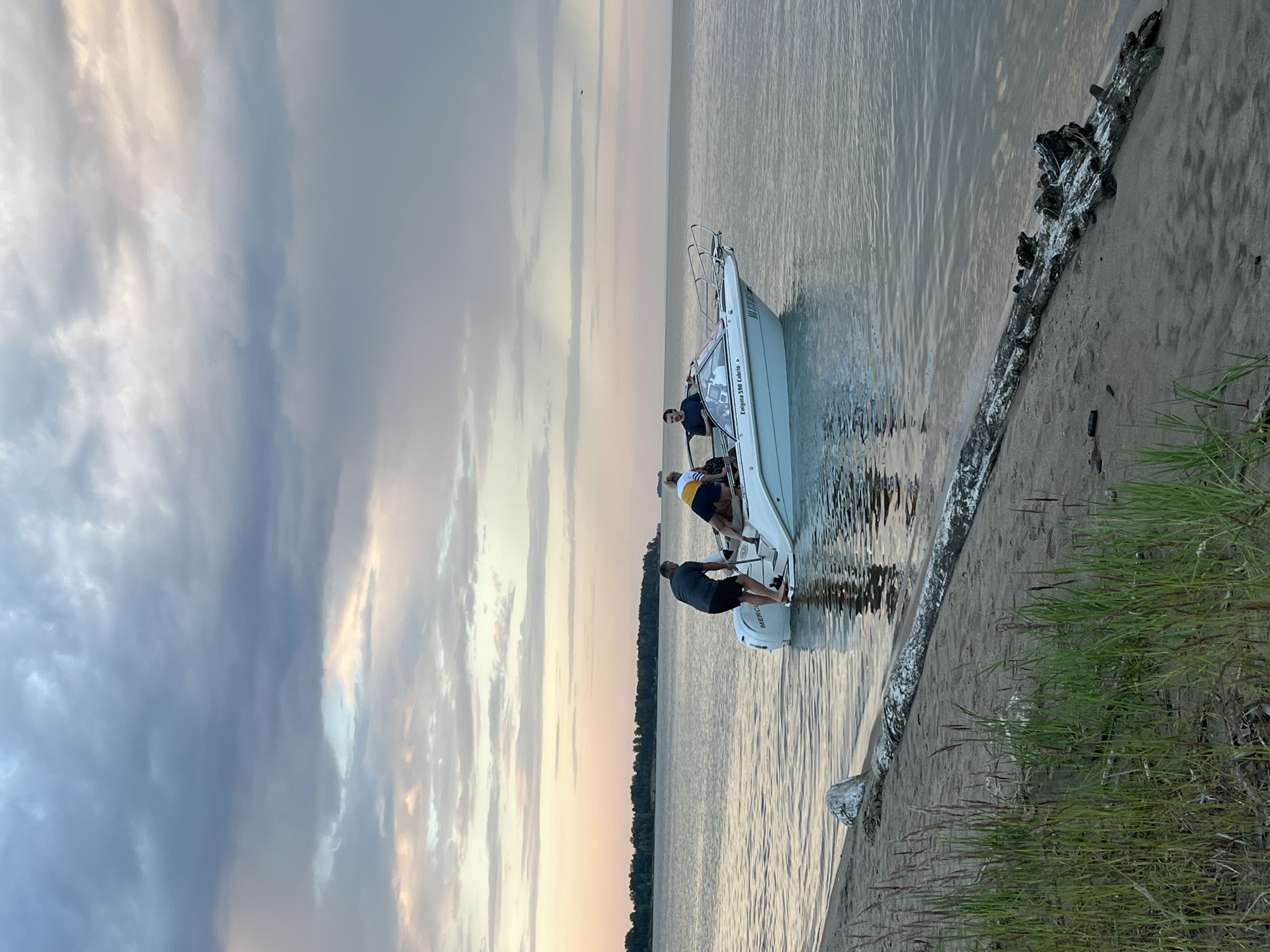 A white motorboat is partially beached near a sandy shore under a cloudy, overcast sky. Three individuals are visible on the boat. One person, wearing a dark t-shirt and shorts, stands near the stern and appears to be pushing the boat. Another person, in a yellow and blue shirt and shorts, is seated on the edge of the stern. A third person, in a dark shirt, is standing near the front of the boat, looking out over the water. The boat has "Enigma 590 Cabrio" written on its side. The water is calm, reflecting the sky. A large piece of driftwood lies on the sand in the foreground, with tall grass growing around it. In the distance, a tree line runs along the horizon. The location is not Borovoe, Russia; this is a generic coastal scene.