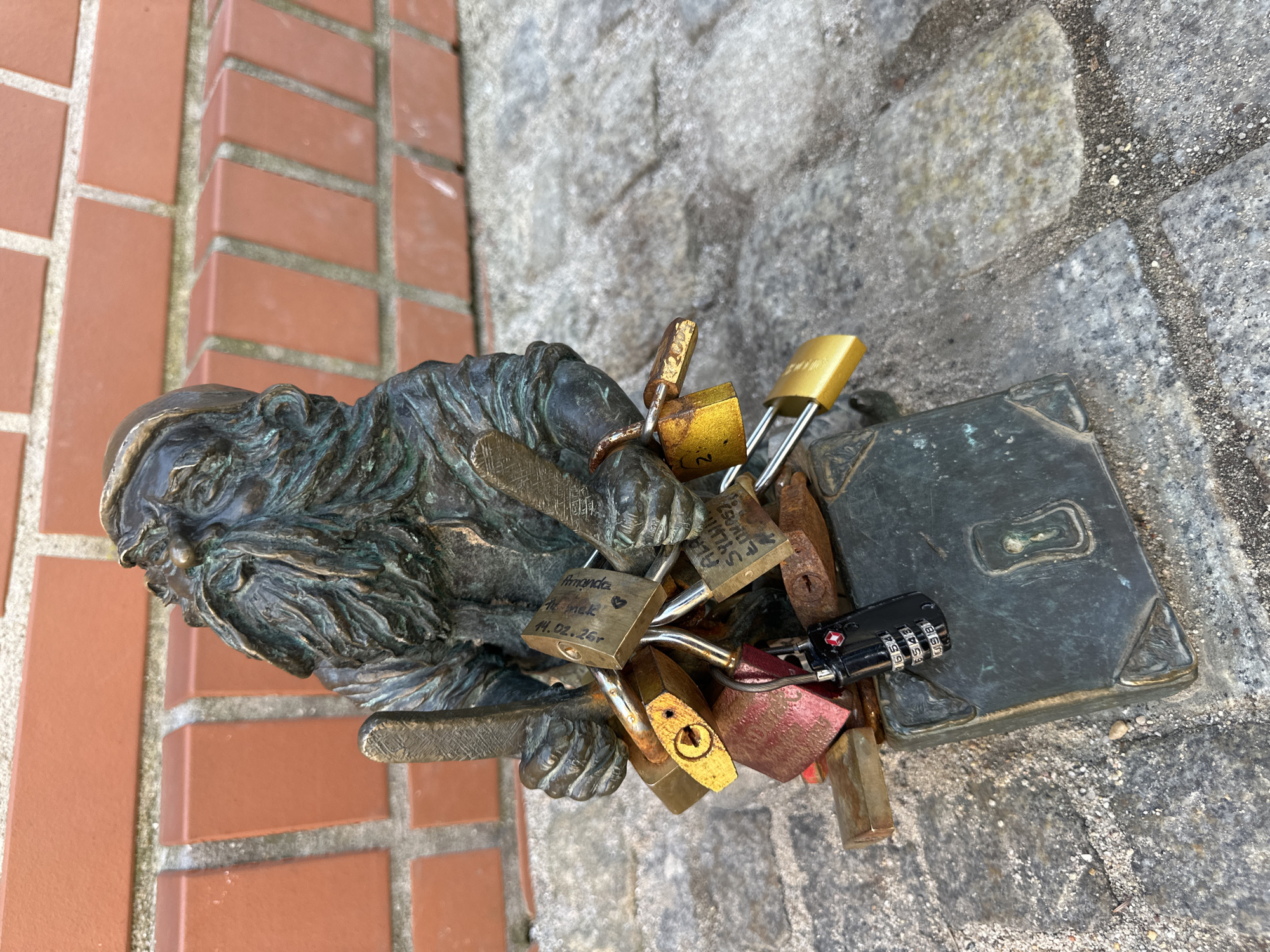 A bronze dwarf statue rests on brick and stone in Wrocław, Poland. The dwarf holds a book-shaped object on which several padlocks are attached. The padlocks are of varying colors and sizes, and some have inscriptions on them. The statue is positioned between a brick wall and a stone surface.