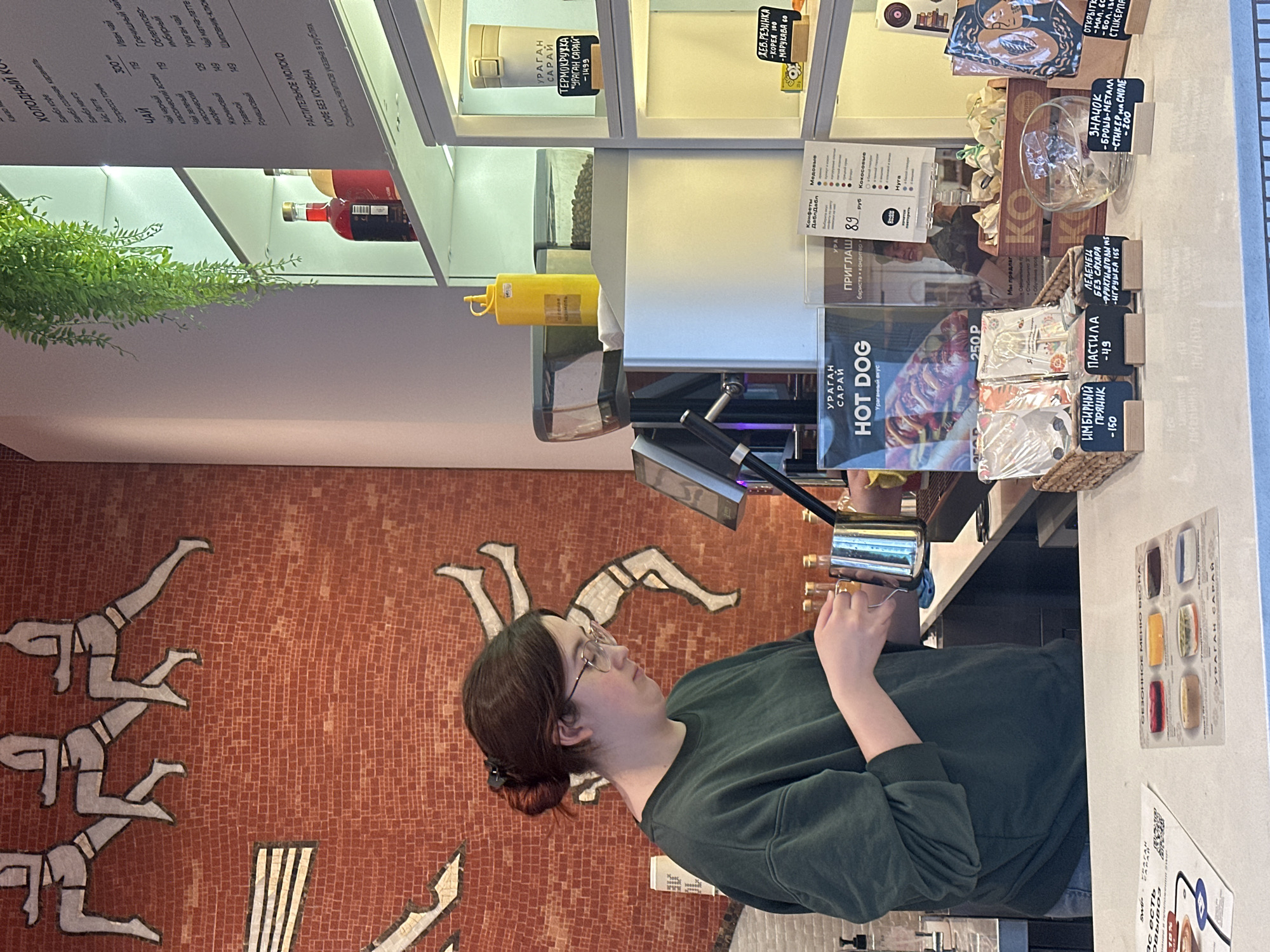 A young woman with glasses is standing at a counter in a cafe. She is wearing a dark green sweatshirt. The cafe has a mosaic wall depicting human figures, and the counter displays a menu board, a hot dog advertisement, confectionery, and coffee. A shelf above the counter showcases food and drink items. The text on the menu is in Russian, and it mentions items related to "URAGAN SARAY." This location is Kazan, Russia.