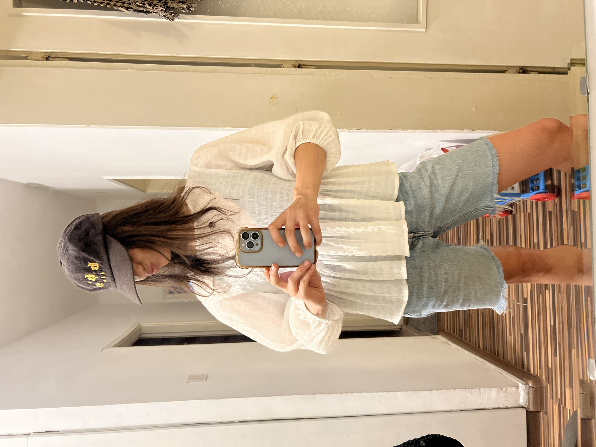 A young woman stands in what appears to be a residential hallway, taking a mirror selfie. She is wearing a dark baseball cap with yellow lettering, a white puff-sleeved blouse, and light-wash denim shorts with a frayed hem. She holds a light blue smartphone in her right hand, using it to capture the reflection. The hallway has light-colored walls and a wood-patterned floor. To the left, a doorway is visible, and to the right, a white doorframe and a portion of a textured wall. A child's toy, partially visible, is on the floor behind her. The image does not provide any specific indicators of its location within the city of Sofia, Bulgaria.