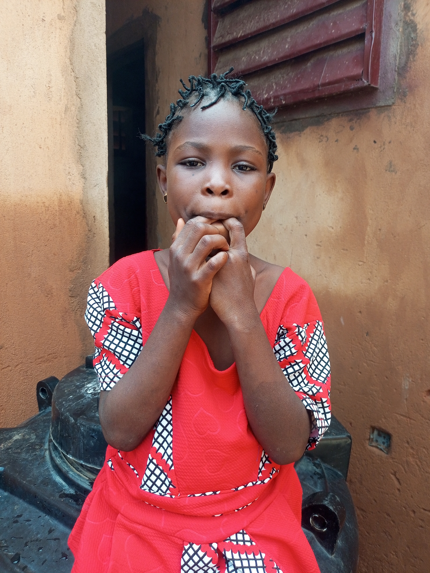 A young girl, with braided hair and a gold hoop earring in her left ear, sits in front of a brown, textured wall. She wears a red dress with a black and white patterned trim. Her hands are raised to her mouth, and she appears to be biting her nails. The background shows a doorway to the left and a window with red louvers above and to the right. A large, black, cylindrical object, possibly a water tank, is visible in the foreground. The scene suggests an outdoor setting, likely in a residential area, possibly in Gombe, Nigeria, given the architectural style of the building.