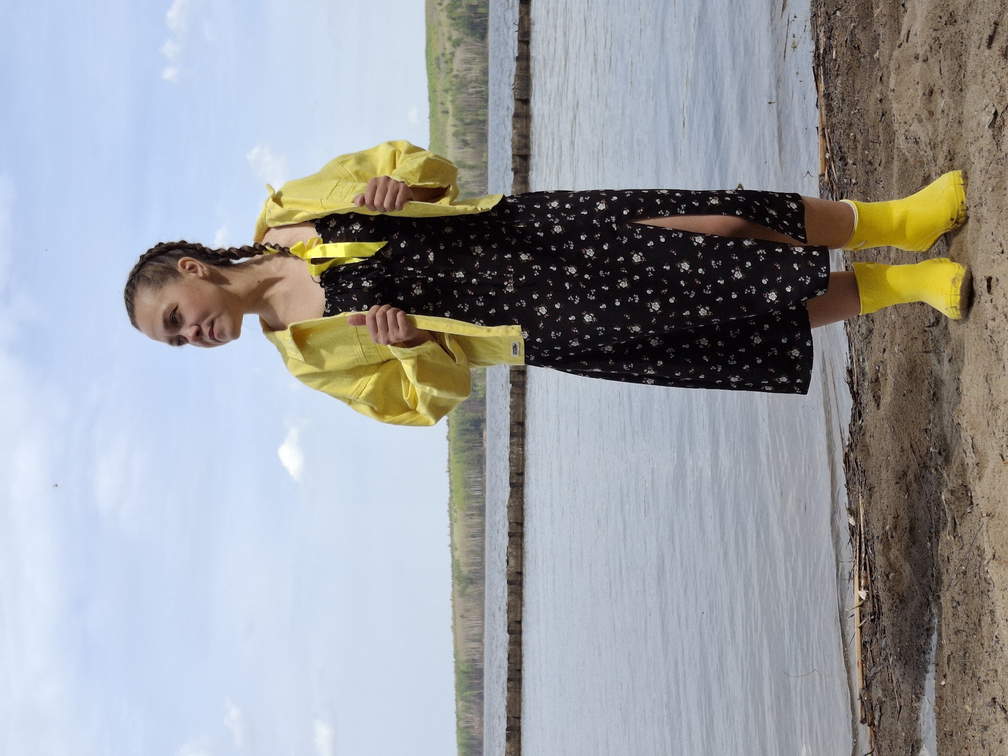 A woman is standing on a beach near the water. She is wearing a black dress with small white flowers, a yellow jacket, and yellow boots. Her hair is braided. The water is calm and there is a blue sky with some clouds. In the background, there are trees on the opposite shore. The location is Chaikovsky, Russia.