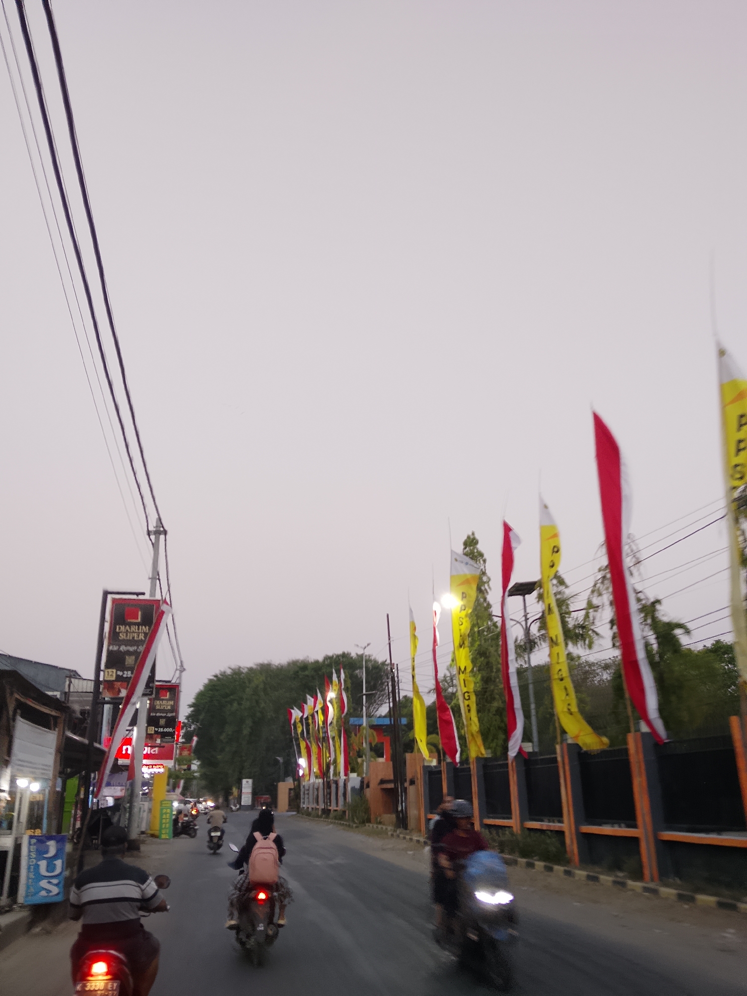 The image shows a street scene in the city of Ngelo, Indonesia. The sky is overcast and a pale gray. Several power lines are visible in the upper left quadrant of the image. Along the right side of the street, a series of red and white Indonesian flags and yellow flags with text are displayed, fluttering in the breeze. Trees and some buildings are visible behind the flags.
On the left side of the street, there are storefronts, including one with a sign for "DIARUM SUPER." Traffic is present on the road. In the foreground, a person is riding a scooter, facing away from the camera. Another person on a scooter is visible further down the road, also facing away. Two more scooters are approaching from the distance, with riders on each. The street is paved and has a curb on the right side. The overall impression is of a moderately busy street during the day.