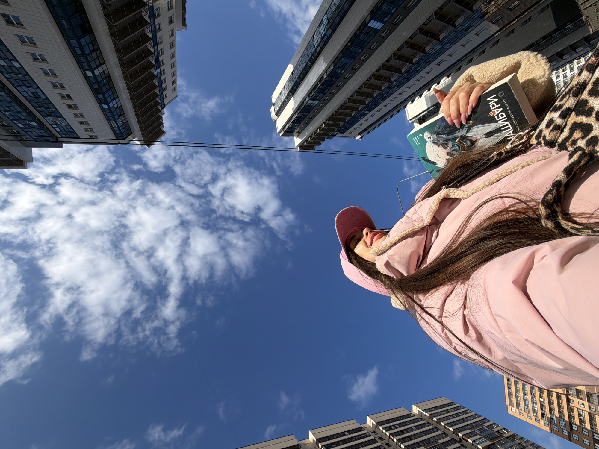 The image depicts an upward-angled view of a female individual holding a book in a modern urban district of Saint Petersburg, Russia. The individual, positioned in the lower-left, wears a pink puffer jacket with a light beige sherpa lining and a matching pink cap. Her long, dark brown hair extends from under the cap. Her face, seen in profile, features red lipstick and a slight smile as she looks towards the upper-right. Her right hand, with manicured nails (pink French tips), holds a paperback book with a dark cover displaying the Cyrillic title "ХАЙШИБУРИ" (HAISHIBURI) and "СНЕГ ПОСЛЕДНЕЙ ЗИМЫ" (Snow of the Last Winter) by V. Sivukha, alongside an illustrative depiction of a stylized face with horns and glowing eyes. A leopard-print bag strap is visible across her torso.
The background consists of several modern high-rise buildings under a clear blue sky with scattered white clouds. To the left, a light-colored multi-story building with numerous windows stands adjacent to a shorter, reddish-brown building. To the right, a tall, contemporary building with a white facade and extensive dark-tinted glass sections, including prominent balconies, dominates the skyline. Two thin black wires traverse the upper sky diagonally. The architecture indicates a newly developed or redeveloped area within the city.