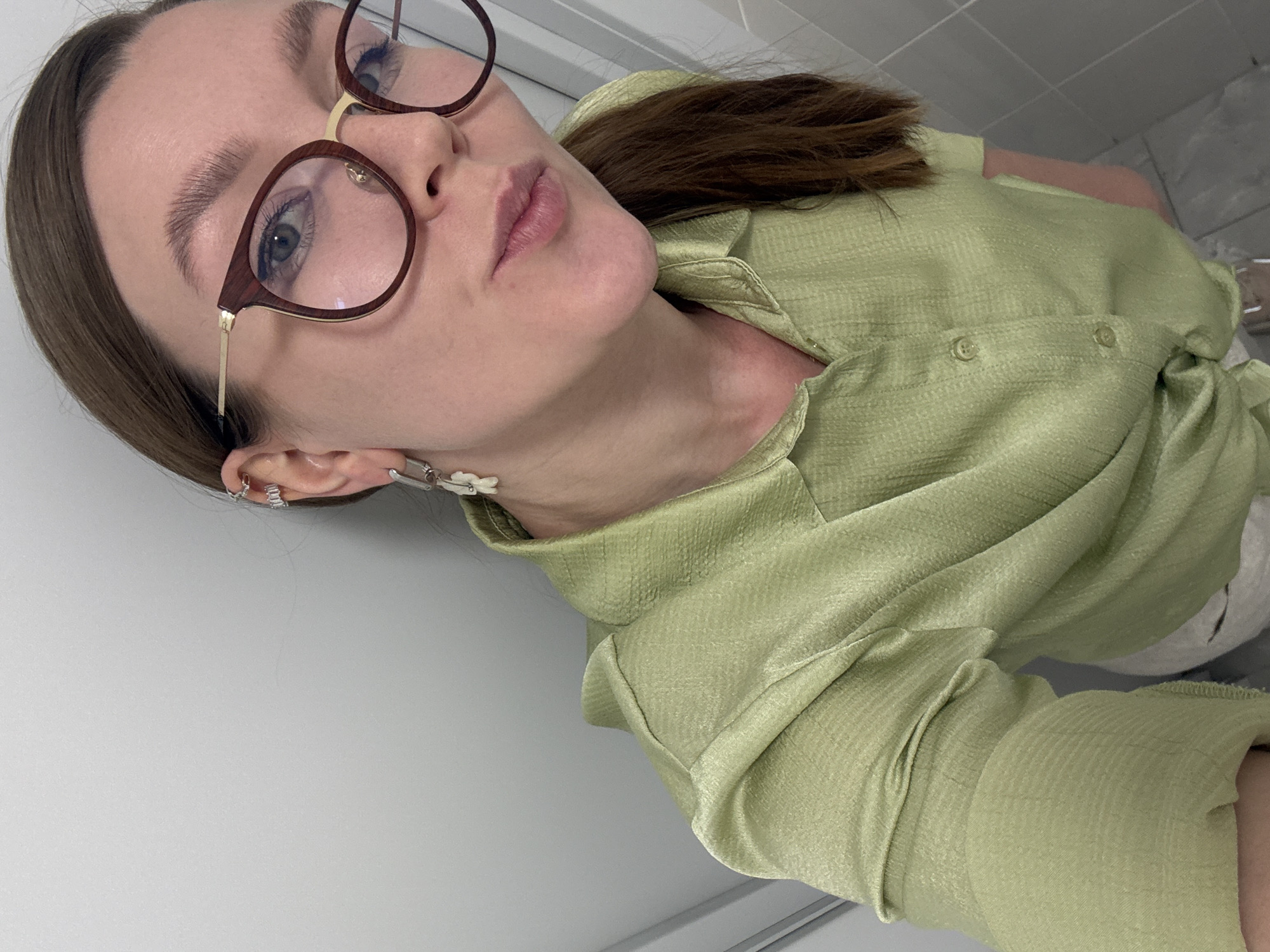 A woman with light skin, brown hair, and glasses is angled towards the camera. She is wearing a light green shirt. The background includes a white wall and a glimpse of a gray tiled floor. The location is unknown and therefore cannot be identified as Izhevsk, Russia.