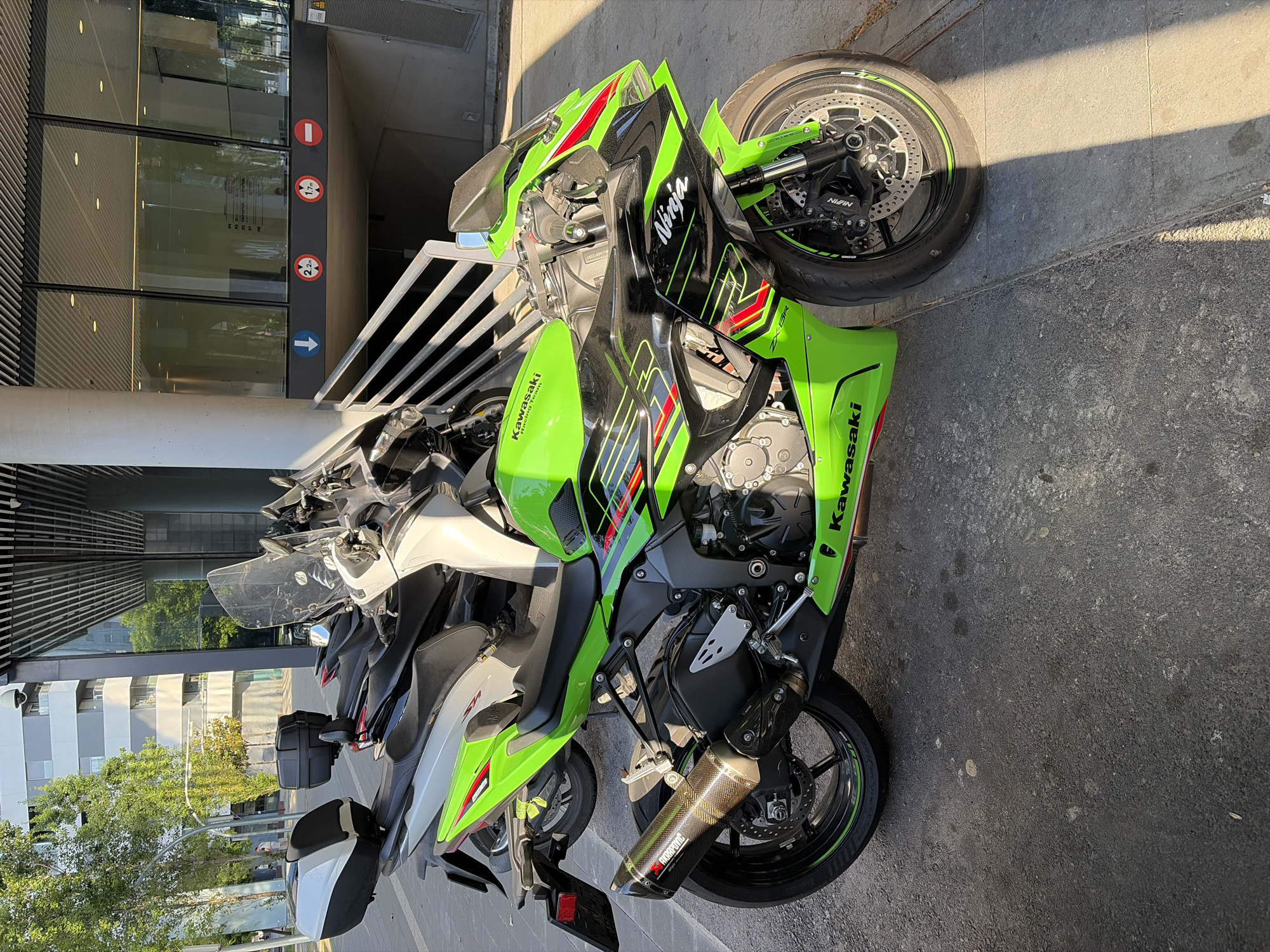 The image depicts a Kawasaki Ninja ZX-10R motorcycle parked in L'Hospitalet de Llobregat, Spain. The motorcycle is green, black, and red, with a "Ninja" decal on the fairing and an Akrapovic exhaust. It is parked next to another motorcycle, which is light-colored. The background includes a modern building with glass windows and metal beams. A traffic sign indicates height restrictions, visible near the building's entrance. The ground is concrete, and sunlight casts shadows across the scene.