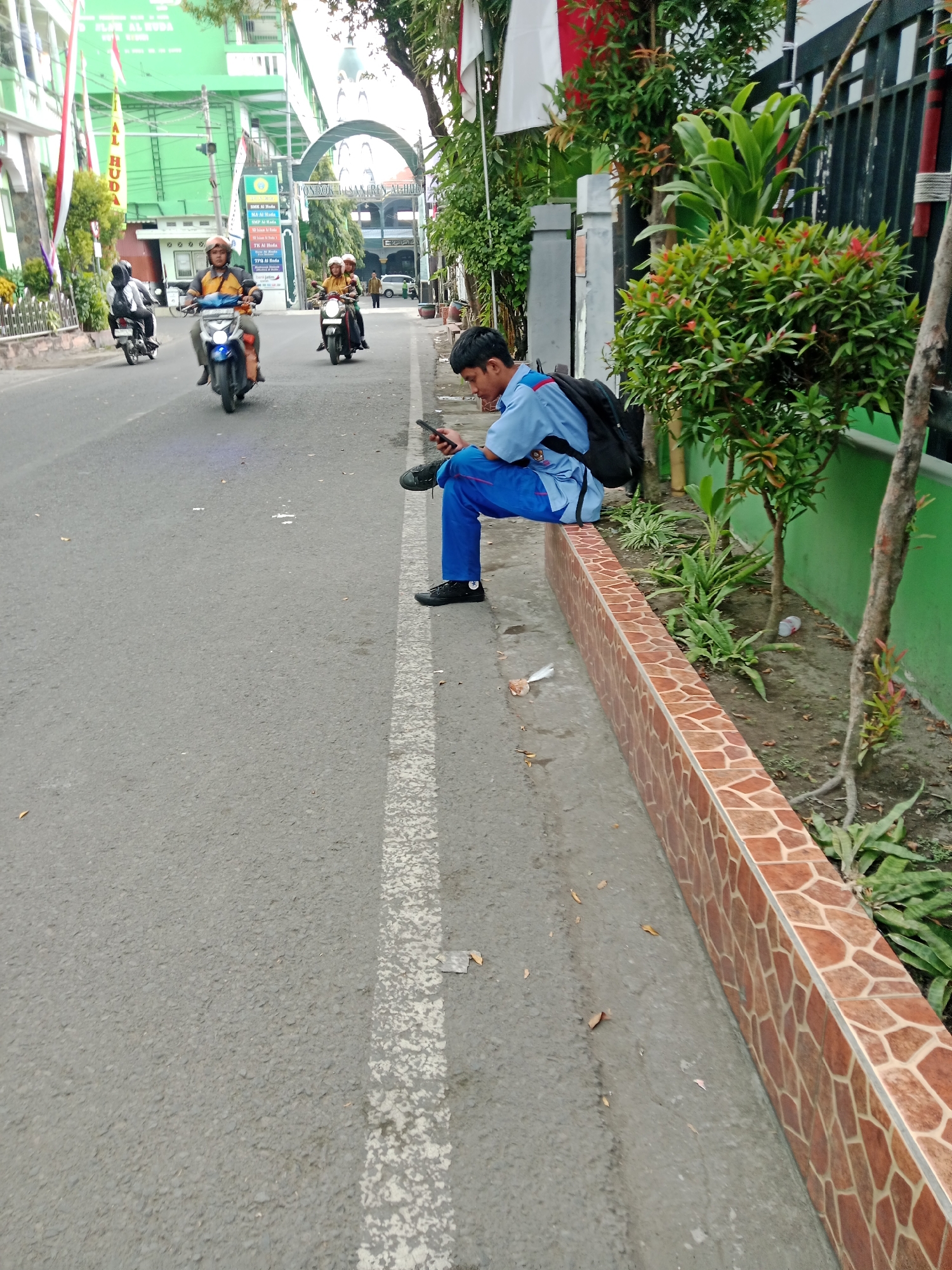 This image captures a street scene in Kediri City, Indonesia, focusing on a young man in a school uniform sitting on a tiled wall beside a road. He is wearing a blue shirt and blue pants, with a black backpack on his back, and is looking at his mobile phone. The road is paved and has a white dashed line marking. Several motorcycles are visible on the road, some with riders wearing helmets, indicating traffic. In the background, there are buildings, a gate with an arch, and trees. The overall atmosphere suggests a typical day in an urban environment within Indonesia.