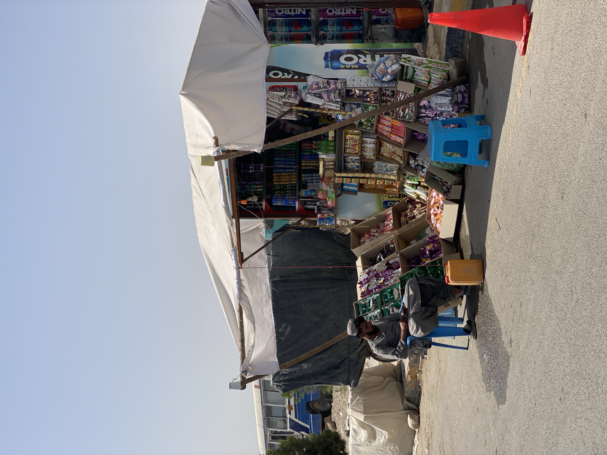 This image captures a street scene in Kandahar, Afghanistan, under a clear, hazy sky. In the foreground, a man in traditional gray clothing and a cap sits on a blue plastic stool, looking down at his phone. He is positioned next to a small stall displaying a variety of packaged snacks and drinks. The stall is covered by a white canvas awning supported by wooden poles. Behind the man, to the left, a blue and white bus is parked on a dusty incline. The ground is paved asphalt, with a puddle reflecting the sunlight. A bright orange traffic cone stands on the right side of the road. The overall environment suggests a roadside market or rest stop, common in urban and semi-urban areas of Afghanistan.