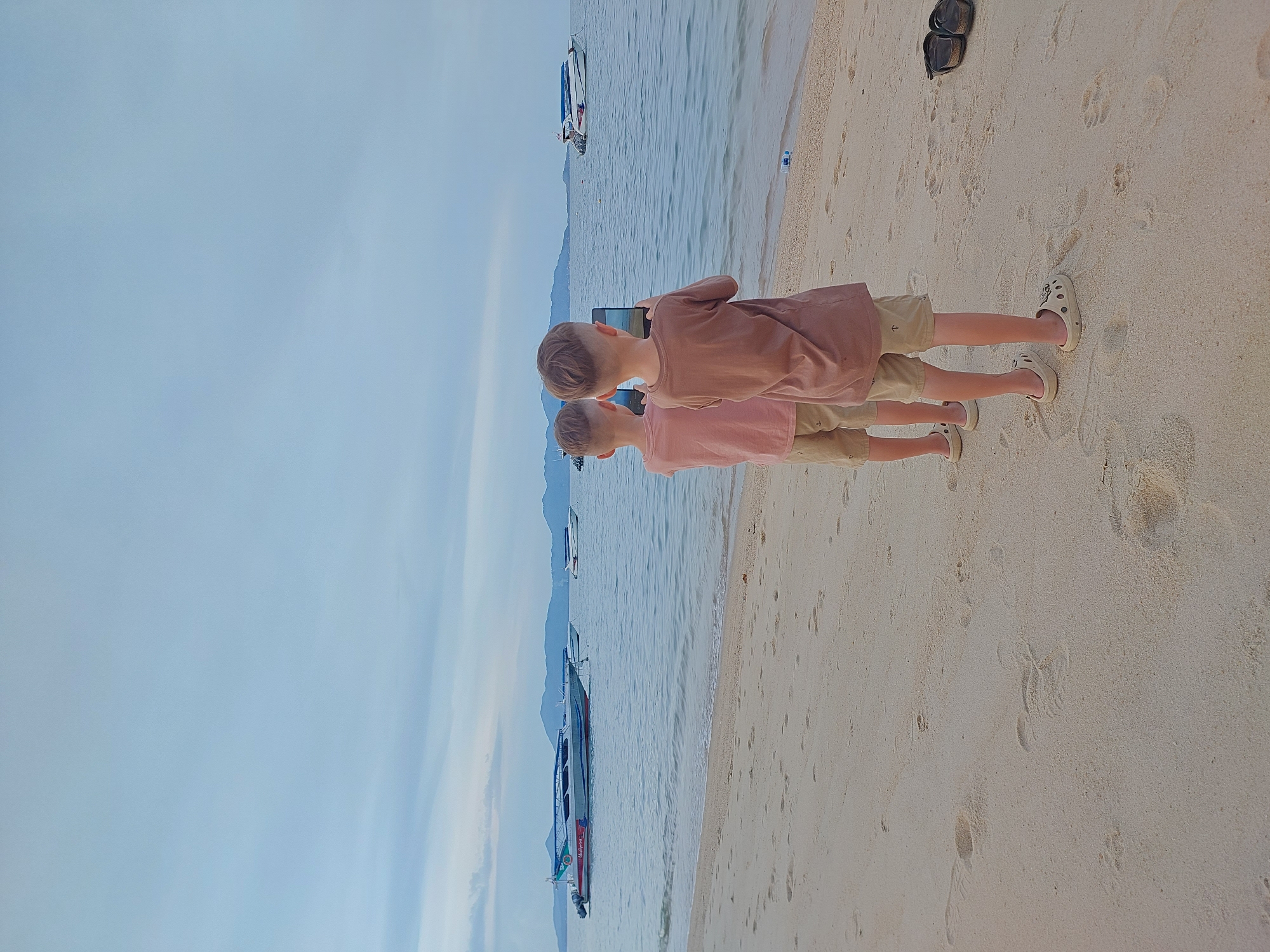 Two young boys, dressed in t-shirts and shorts, stand on a sandy beach in Baan Tai, Thailand, facing the ocean and holding up mobile phones to take pictures. The boy on the left wears a pink t-shirt and tan shorts, while the boy on the right wears a brown t-shirt and tan shorts with an anchor detail. Both boys are wearing light-colored clogs. In the background, calm ocean water stretches towards a distant shoreline with mountains. Several boats, including speedboats and a larger boat with a red stripe, are visible on the water. The sky is overcast with pale blue and grey clouds. The sand on the beach is light in color and shows numerous footprints. A pair of dark sandals lies on the sand to the right of the boys.