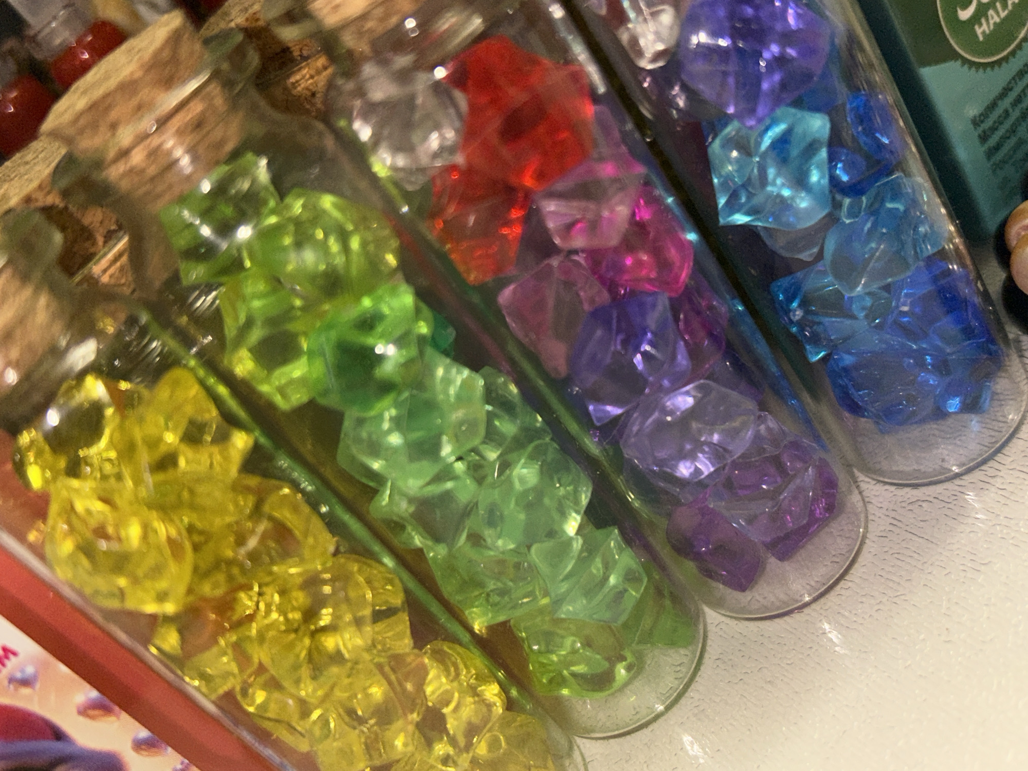 The image shows several glass vials filled with colorful, translucent, crystal-like objects. The vials are arranged diagonally across the frame, with their cork stoppers visible at the top. From top to bottom, the colors of the crystals appear to be yellow, green, red and pink, purple, and blue. The crystals are irregularly shaped and catch the light, creating a sparkling effect. The vials are placed on a textured white surface. In the upper left corner, part of a red box and a glimpse of a patterned surface are visible. In the lower right corner, a portion of green packaging with some text in a foreign language, and a circular green and white seal is visible. The scene appears to be an indoor setting, possibly a retail display or a collection of decorative items. There are no people or discernible actions taking place. The overall impression is one of vibrant color and texture.