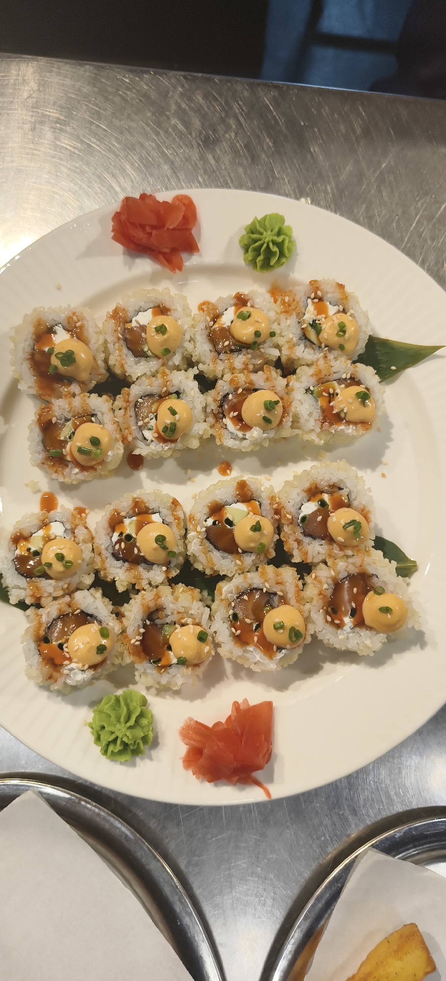 On a stainless steel surface in an unknown city and unknown location, a white oval plate is filled with sushi rolls, arranged in two rows. The sushi rolls appear to be of the same variety, featuring rice on the outside, sprinkled with sesame seeds, and topped with a creamy orange sauce and a small green herb garnish. Inside the rolls are darker fillings, possibly fish or vegetables, and a creamy white substance. The plate also contains small piles of pickled ginger (reddish-orange) and wasabi (green) in opposite corners. The scene suggests a culinary setting, likely a restaurant or food preparation area. Part of two metal trays with white paper lining are visible in the lower portion of the frame.