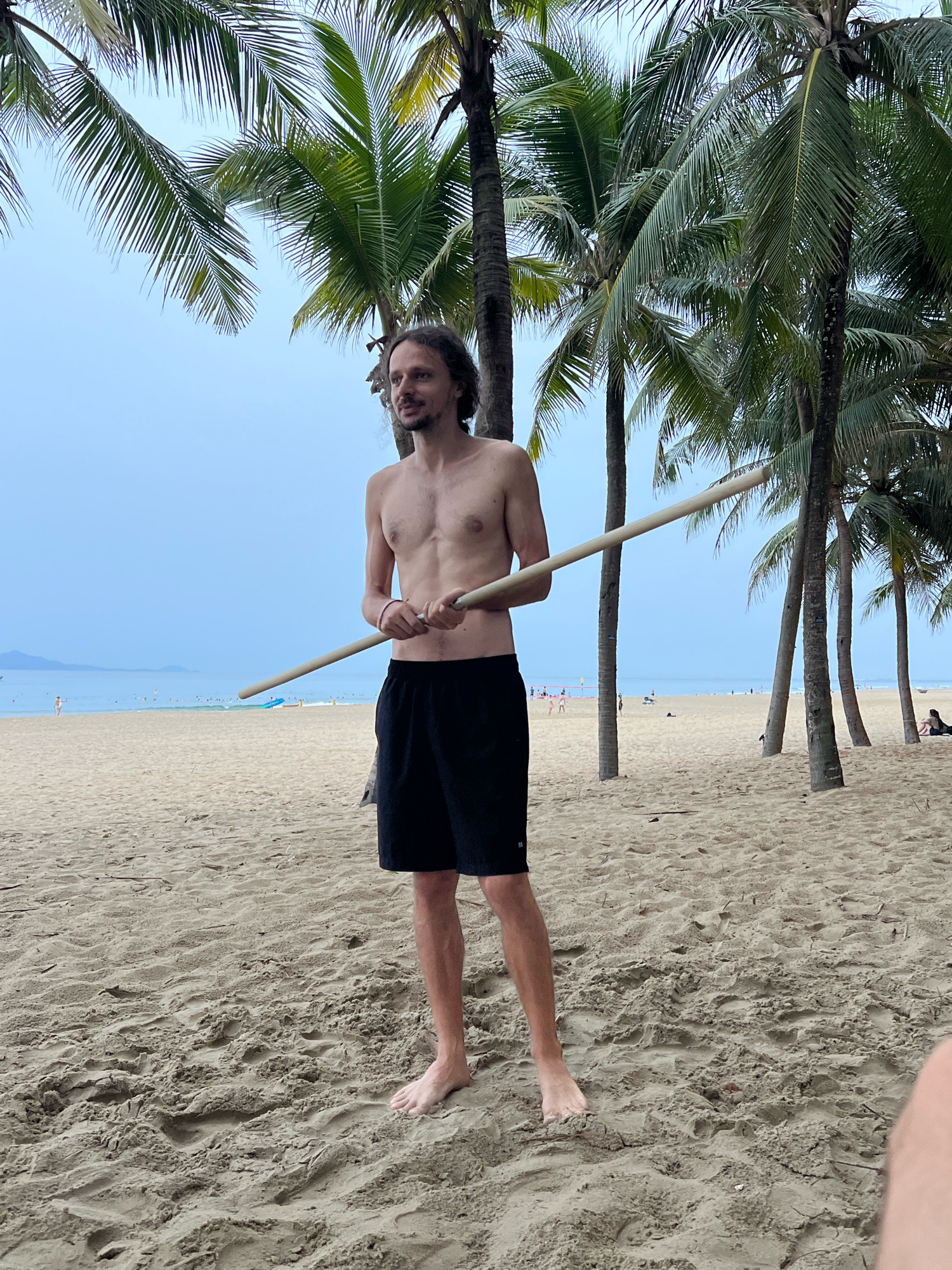 A man stands on a sandy beach in Da Nang, Vietnam, holding a long wooden pole horizontally with both hands. He is wearing black shorts and is shirtless, revealing his torso. The background shows palm trees, the ocean with several boats and people, and a hazy sky. The sand is textured with footprints and debris.