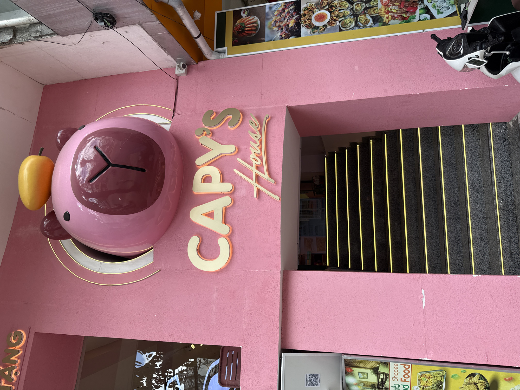 The image shows the exterior of a pink building with a large, stylized pink bear's head as a prominent architectural feature. Above the entrance, which features a set of dark stairs with yellow accent lighting, is a neon sign that reads "CAPY'S House". The building is located in Nha Trang, Vietnam, as indicated by the signage "TANG" at the top left of the building and a sign featuring the Shopee Food logo on the left side. A motorcycle is parked in front of the building on the right side. The building's facade is textured, and there are advertisements for food displayed vertically on the right side of the entrance. The scene appears to be on a street in a commercial area.