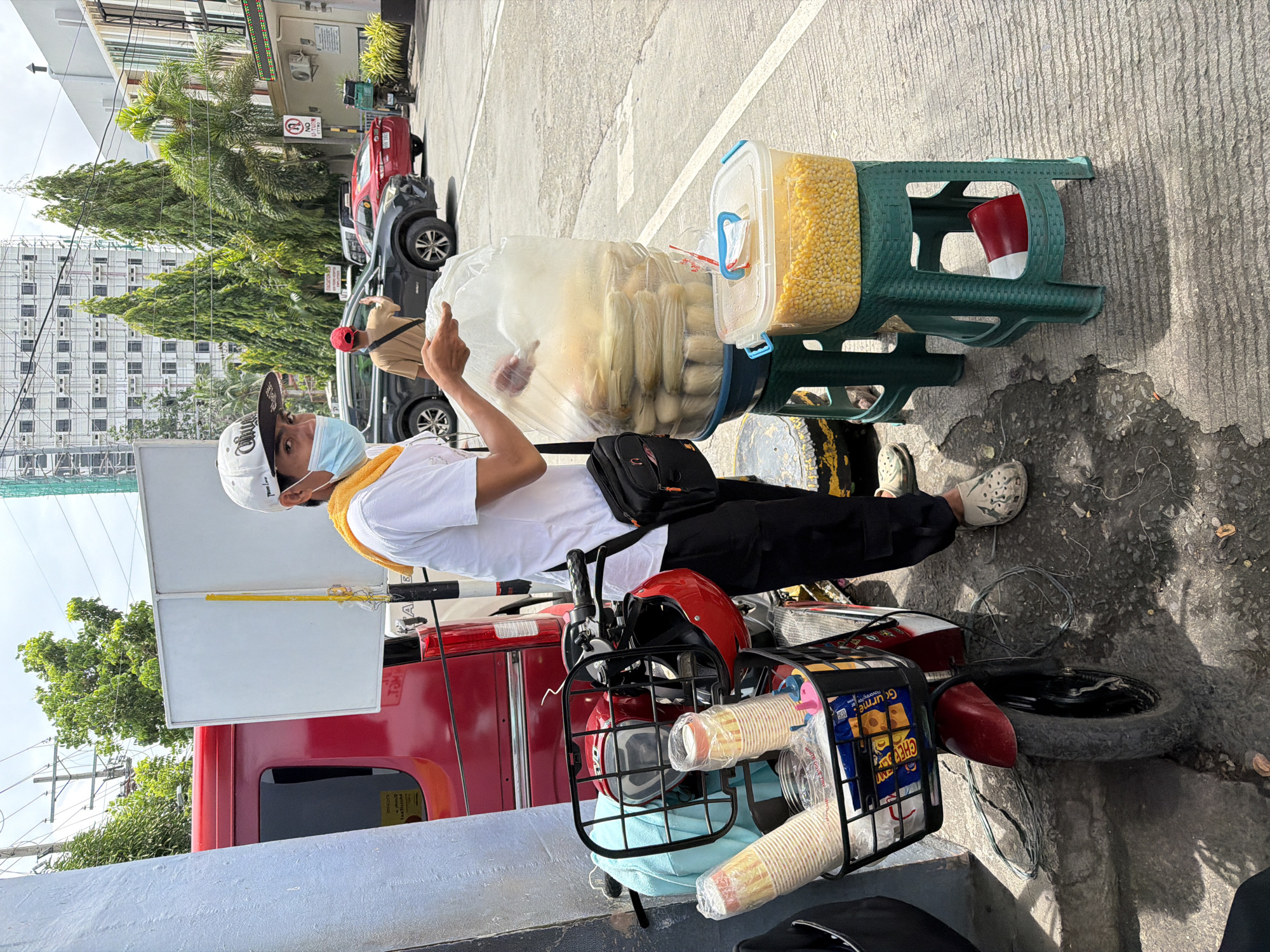 A young man, wearing a white t-shirt, black pants, a white baseball cap, and a light blue face mask, stands on a street in Davao City, Philippines. He is holding a large, clear plastic bag filled with corn on the cob, and appears to be serving it. Next to him, on a green plastic stool, is a clear plastic container filled with loose corn kernels. A red scooter is parked beside him, with a red helmet in the front basket along with paper cups and a package of snacks. In the background, a red truck, several cars, and buildings can be seen. The street is paved with asphalt, and there are trees and overhead wires in the scene. The overall atmosphere is that of a street vendor operating outdoors.