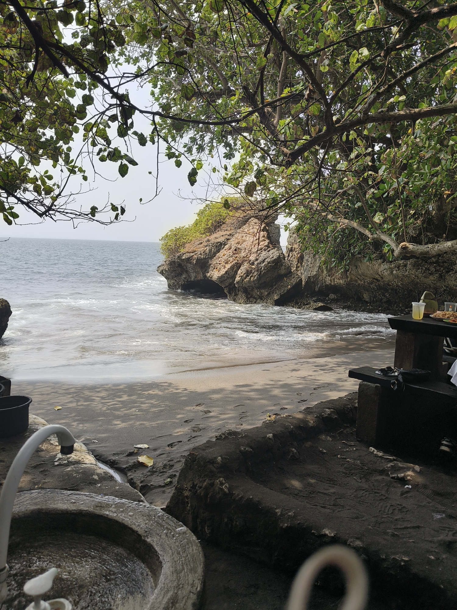 The image depicts a secluded beach area with a view of the ocean and rocky cliffs. In the foreground, a stone sink with a faucet is visible, along with a black bucket. Further back, a wooden table and bench are set up, with a drink and some food items on the table. The beach itself is sandy with some scattered leaves and footprints. Lush green trees overhang the scene from above, casting shadows on the sand. The ocean is calm with small waves breaking on the shore. The overall impression is one of a peaceful, natural setting, possibly a picnic or relaxation spot. The location is identified as Desa Batukaras, Indonesia.