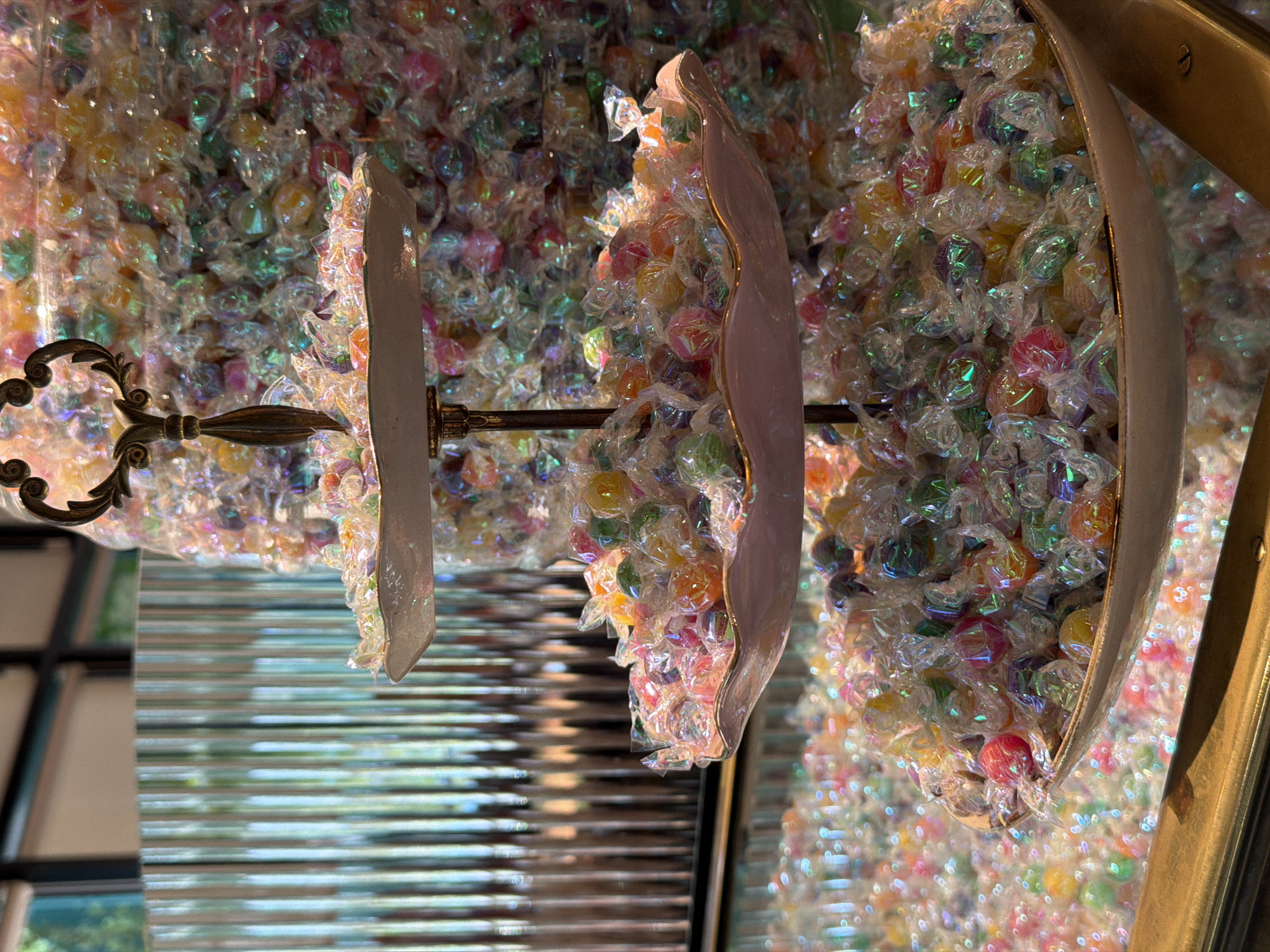 The image shows a chandelier made of iridescent wrapped candies. The chandelier has three tiers, with a metal frame and ornate details. It is reflecting in a mirror, which also shows a window with horizontal blinds. The location appears to be in the city of Tel-Aviv, Israel.