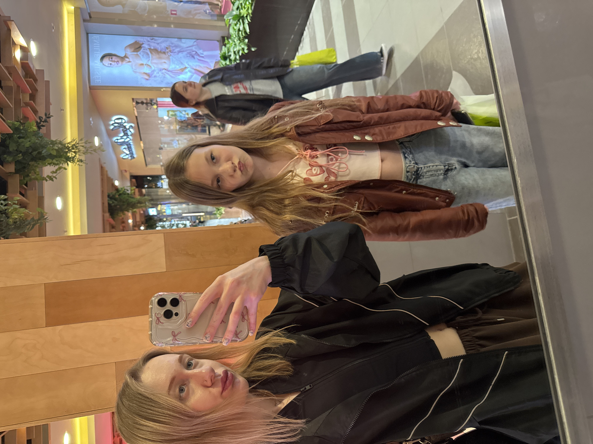 A selfie is taken in front of a mirror, reflecting three people. The foreground shows a woman holding an iPhone with a clear case decorated with bows. She wears a black jacket with white stripes and has blonde hair. The reflection shows a younger girl with blonde hair, wearing a brown jacket, a pink top, and blue jeans. Further back in the reflection is a person wearing a leather jacket and blue jeans. The location appears to be a shopping mall in Ol'ginskaya, Russia, based on signage and architectural style in the background. The environment includes storefronts, displays, and greenery. The interaction is a group taking a picture of themselves in a mirror at a shopping center.