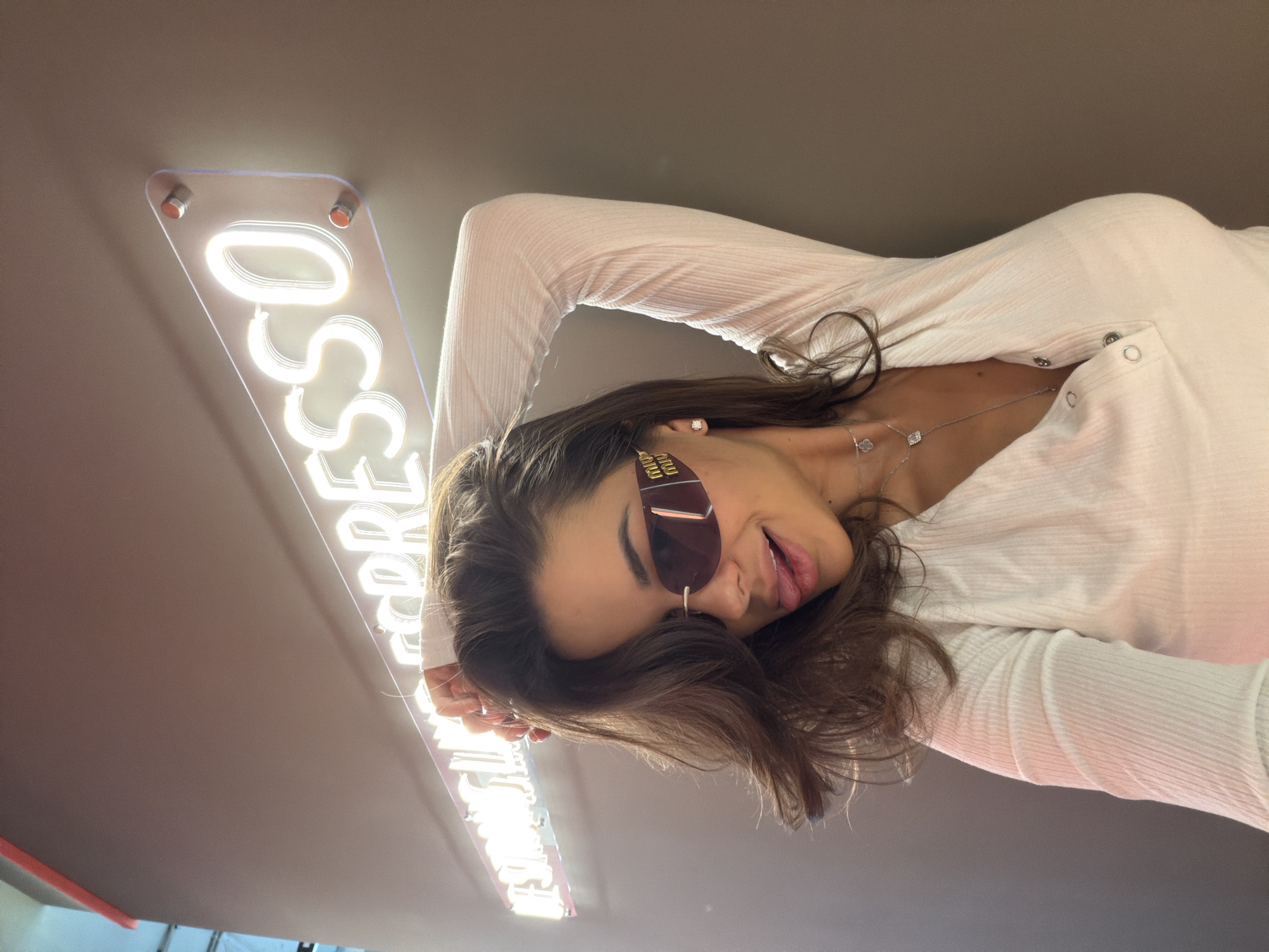 A young woman wearing sunglasses and a white long-sleeve shirt poses in front of an "Espresso" sign. The sign is lit with white neon and appears to be mounted on a ceiling or high wall. Based on the sign and general aesthetic, this is likely a cafe. While the city is not discernible from the image, the prompt specifies Krasnoyarsk, Russia, as the city. The woman is close to the camera, with her head tilted. She has dark hair and is wearing a necklace. The environment appears to be indoors with a neutral-toned ceiling.
