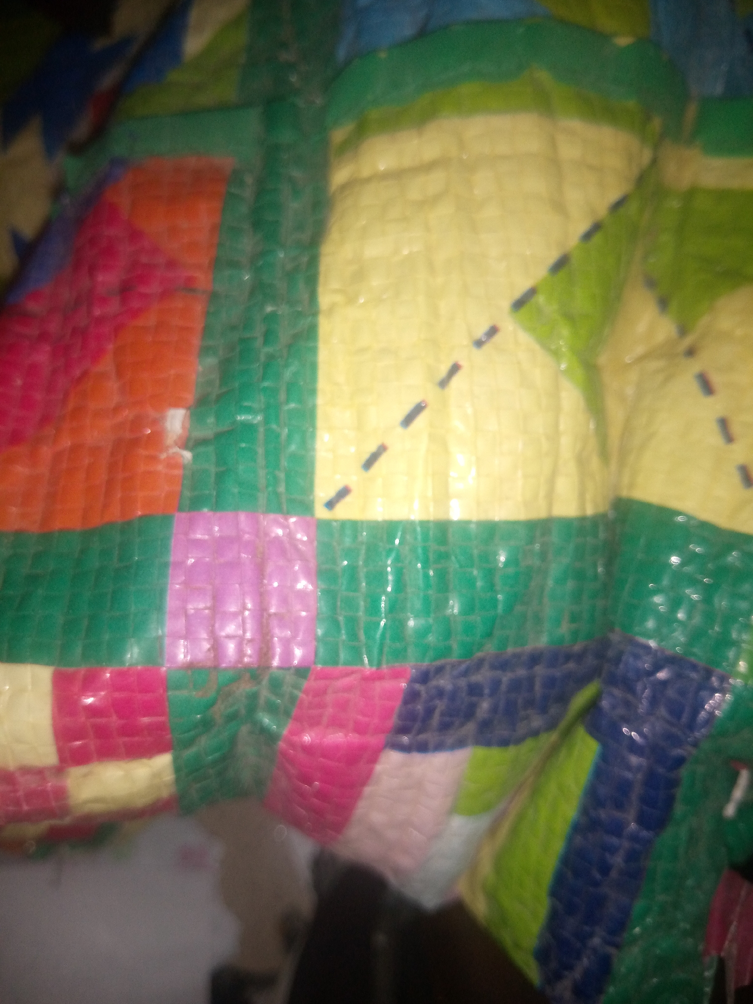 A colorful, quilted object is displayed in close-up against a blurred background. The object is made of various colored rectangular panels, including pink, orange, yellow, green, and blue, arranged in a patchwork pattern. Some panels have subtle textures, and there are occasional small, dark dash-like marks on some of the yellow and green sections. The lighting creates reflections on the shiny surface of the material. The overall impression is of a vibrant and somewhat abstract textile. The location is not discernible from the image.