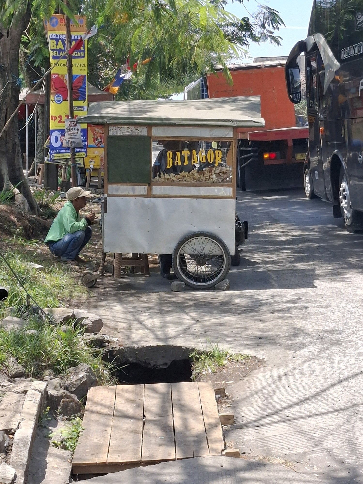 A person is crouching beside a food cart on a sunny day in Sukalarang, Indonesia. The food cart is small, with a metal body and a wooden frame. It has a sign that says "BATAGOR" in yellow letters, and it is filled with what appears to be food items. The person, wearing a green shirt and blue jeans, is looking down at something in their hands. The background shows trees, banners, and vehicles, suggesting a roadside setting. The ground is paved with asphalt, and there is a patch of grass with rocks on the left side of the image. A wooden plank bridge spans a small ditch in the foreground.