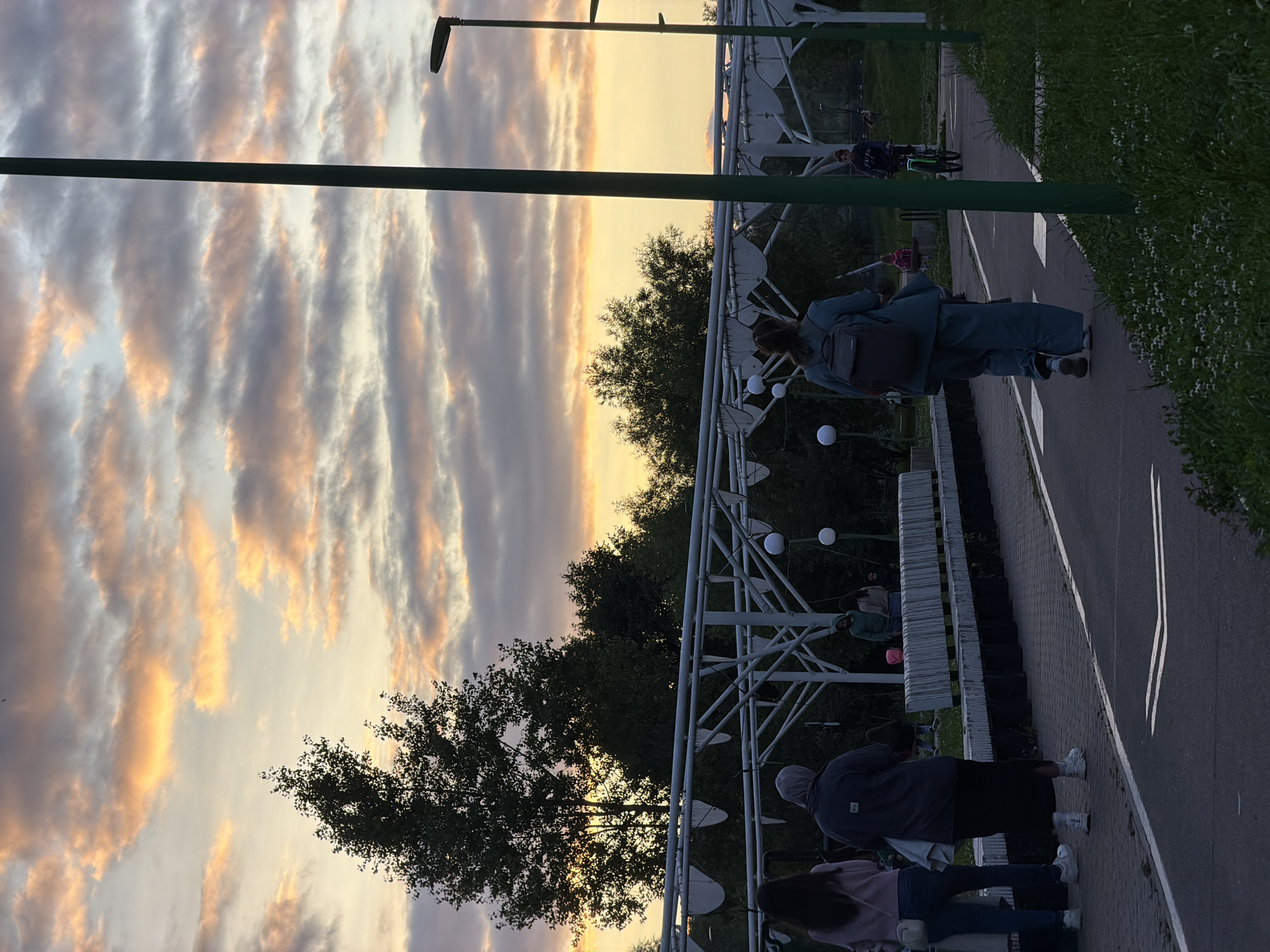 A vertical, eye-level, medium shot captures a park scene at dusk in Kazan, Russia. The sky is filled with dramatic, colorful clouds, painted in hues of orange, yellow, and purple, suggesting either sunrise or sunset. In the foreground, a paved pathway, possibly a bicycle or pedestrian lane, runs diagonally from the bottom right to the center left. A tall, green lamppost stands prominently on the right side of the frame, with another lamppost further in the background.
On the pathway, a person wearing a teal-colored outfit and a backpack is walking away from the camera, towards the left. To the left of this person, on a paved walkway, two individuals are standing, facing away from the camera. One is wearing a pink top and jeans, and the other is wearing a grey hooded jacket and a black skirt. In the mid-ground, a white, modern-looking structure with a lattice-like design stretches horizontally across the image. Behind this structure, lush green trees are visible, silhouetted against the colorful sky. Several people are scattered in the mid-ground, some standing and some appearing to be sitting on benches. In the background, a young person on a bicycle is riding on the pathway. The ground is covered with grass, with small white flowers blooming in the lower right corner. The overall atmosphere is serene and contemplative, with the vibrant sky adding a touch of drama to the scene.