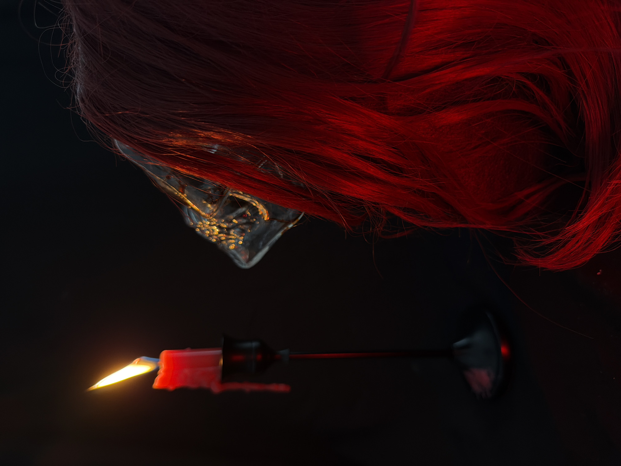 The image depicts a dark, intimate scene with a single red candle burning in a black candlestick holder on the left. The candle flame casts a warm glow. To the right, the back of a person's head is visible, featuring long, vibrant red hair. The person is leaning towards the candle and appears to be holding a clear, faceted glass object close to their face. The background is entirely black, creating a stark contrast and emphasizing the illuminated elements. The overall mood is mysterious and somewhat dramatic, with the interplay of light and shadow. The location is unknown.