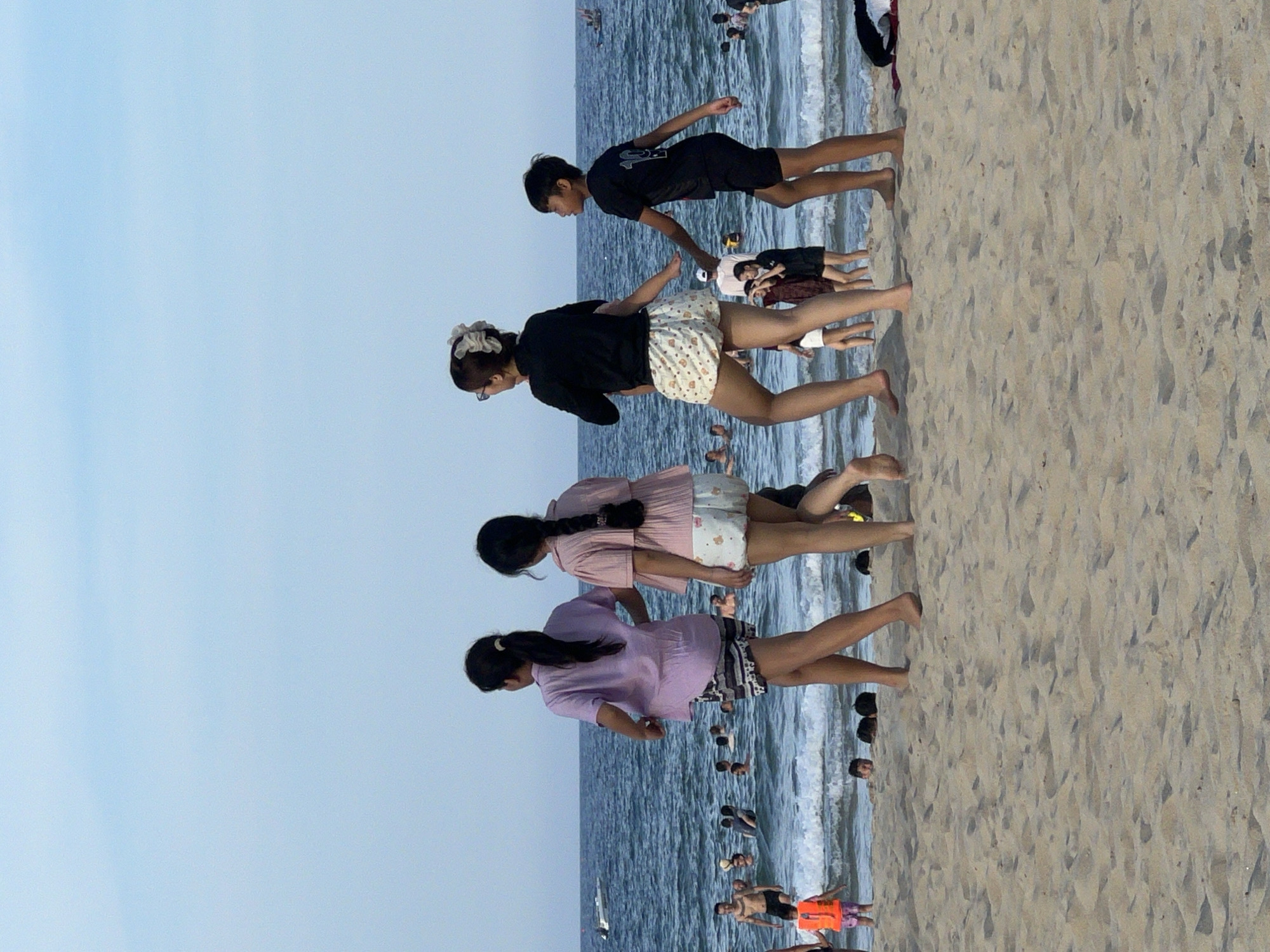 In the city of Đà Nẵng, Vietnam, on a beach with fine sand and a gentle surf, four individuals are walking from left to right in the foreground. A young girl in an orange life vest and a child in pink shorts are further left near the water's edge. In the water, several people are swimming and playing in the waves. The sky is clear and blue.