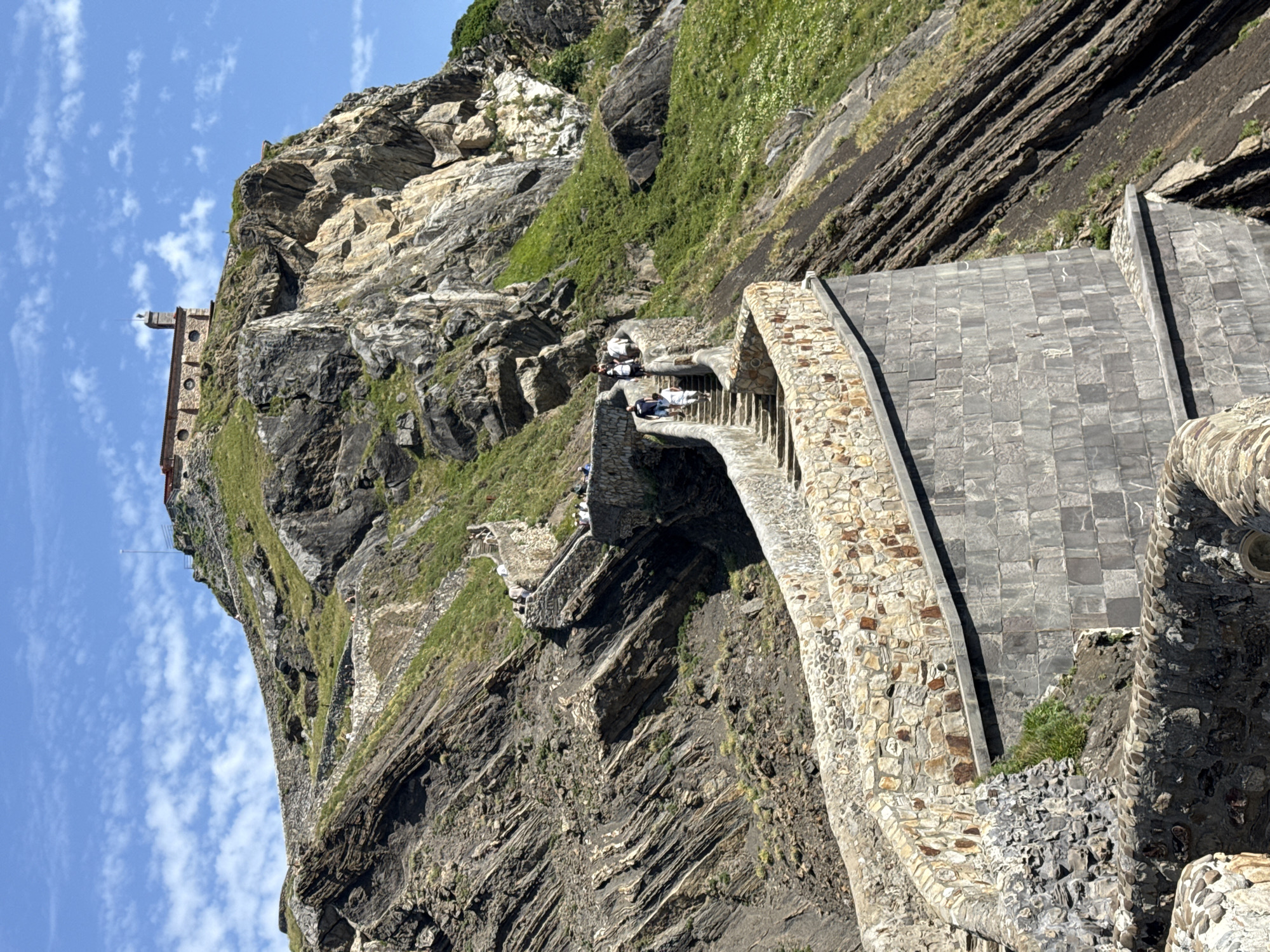 This is a photograph of the San Juan de Gaztelugatxe hermitage, a popular tourist destination located on a small islet off the coast of the Basque Country, near the town of Bakio, Spain. The image captures a dramatic view of a winding stone staircase leading up a steep, rocky hillside towards a small, ancient building perched on the summit.
The staircase is constructed from rough-hewn stones and is lined with stone walls, some of which are reinforced with darker, square-cut stones. The path curves and ascends in a series of switchbacks, offering a challenging but scenic climb. Several people are visible on the stairs, appearing to be tourists walking up towards the hermitage. They are dressed in casual, warm-weather clothing.
The surrounding environment is rugged and natural. The hillside is covered in patches of green grass and sparse vegetation, interspersed with dark, striated rock formations that showcase the geological history of the area. The cliffs are steep and imposing, rising sharply towards the bright blue sky, which is dotted with scattered white clouds.
At the very top of the hill, the hermitage, also known as the hermitage of San Juan, is visible. It is a small, stone building with a red-tiled roof and a prominent bell tower. A small lighthouse is also attached to the building. The structure appears weathered, hinting at its age and exposure to the elements.
The overall impression is one of a historic and picturesque location, combining natural beauty with man-made architecture. The photograph is taken from a low angle, emphasizing the grandeur of the staircase and the challenging ascent to the hermitage. The lighting suggests a sunny day, casting shadows that highlight the textures of the stone and rock.
